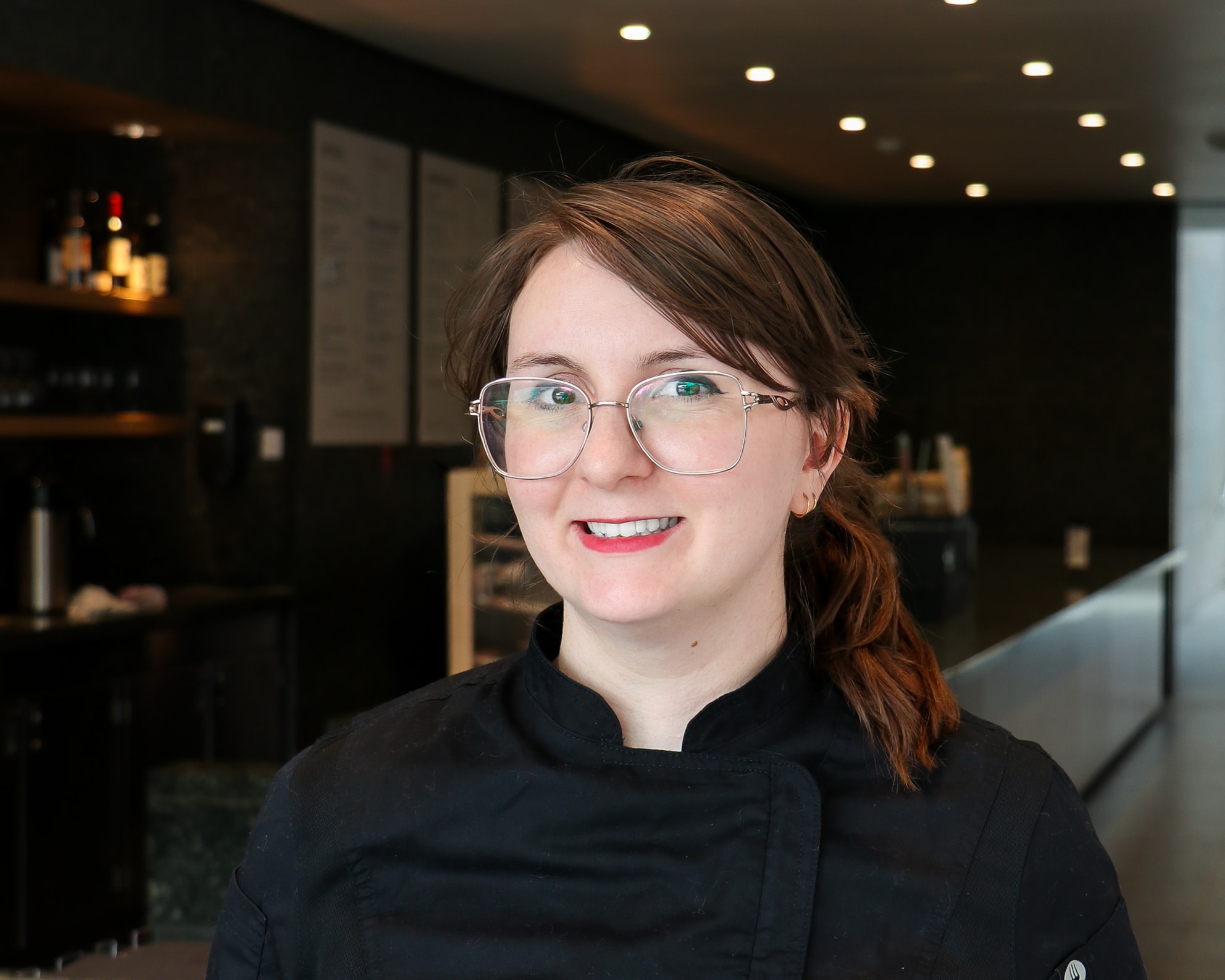 A woman with glasses and brown hair tied back, wearing a black chef’s coat, smiles at the camera. She is standing in a modern, softly lit restaurant or café interior.