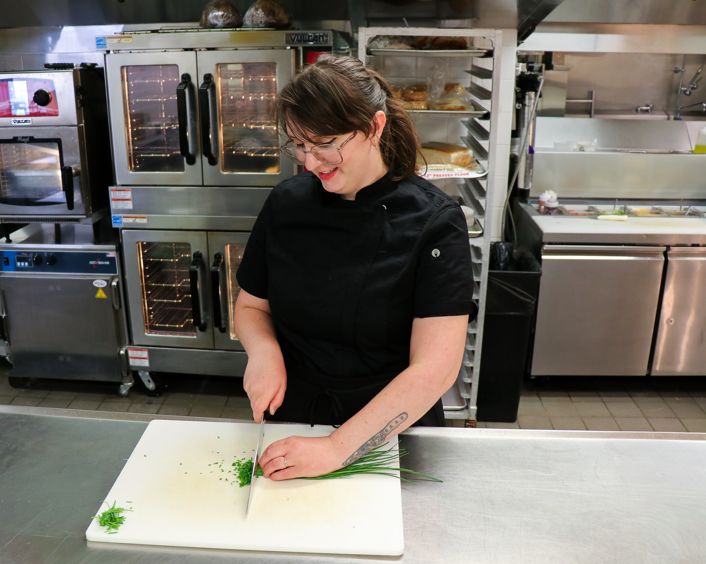 A chef in a black uniform chops fresh green herbs on a white cutting board in a commercial kitchen, smiling while working. Ovens and stainless steel appliances are visible in the background.