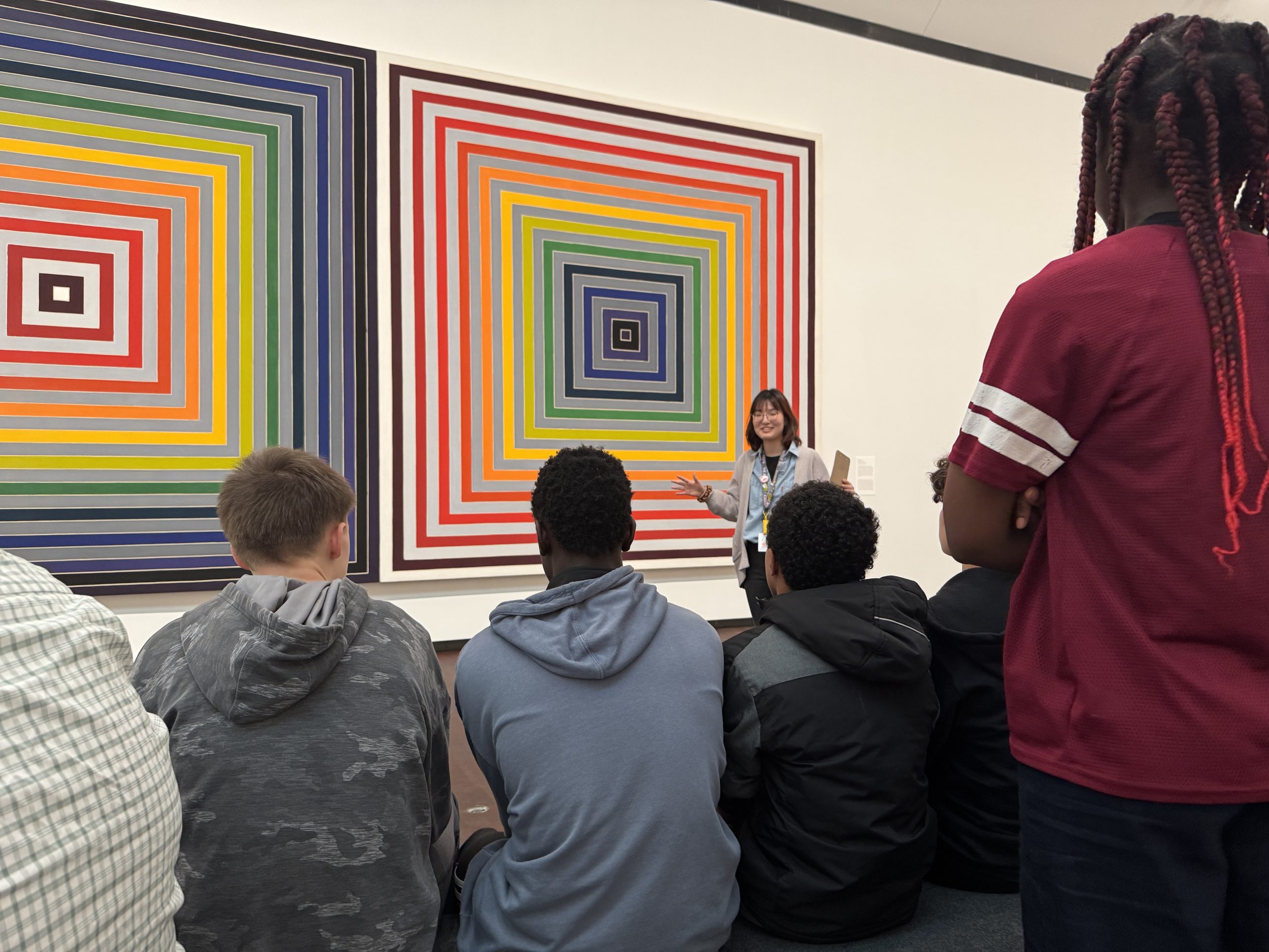 A group of students sit in front of two large, colorful, geometric paintings of concentric squares while a woman stands and speaks, gesturing toward the artwork in a gallery or museum.