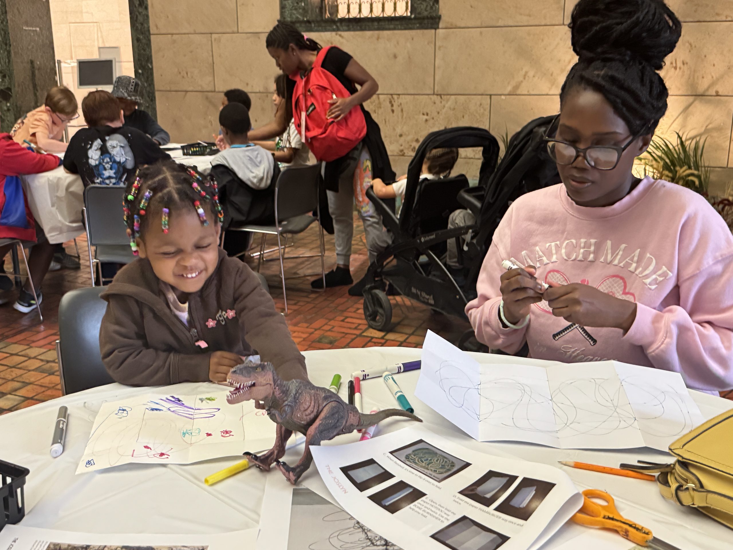A young child and an adult sit at a table drawing with markers. A toy dinosaur and drawing supplies are on the table. The child laughs while the adult colors. Other people work at tables in the background.