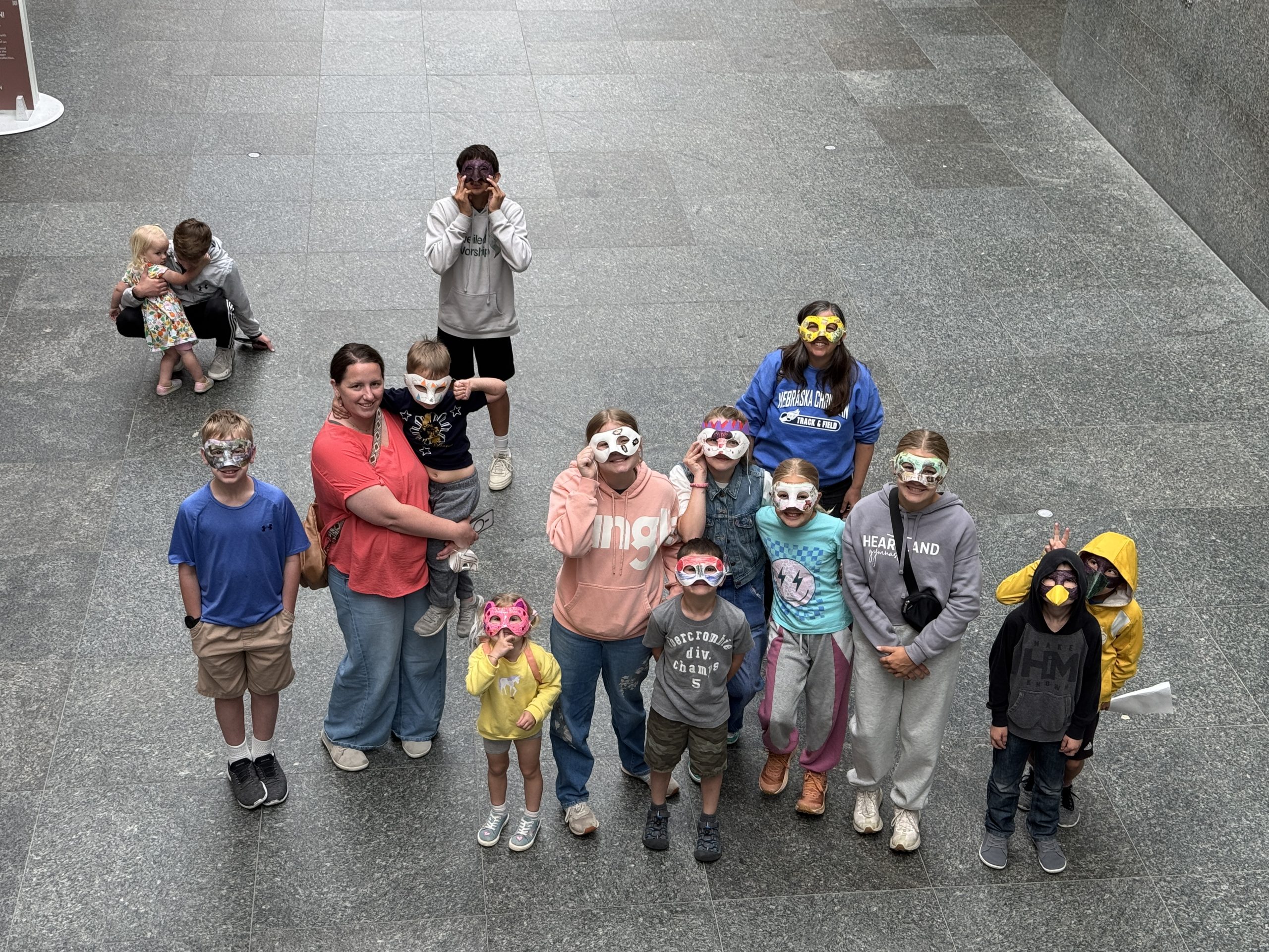 A group of children and adults wearing colorful animal masks stand together indoors on a gray tiled floor, looking up at the camera and posing for a photo. In the background, two adults sit with small children.