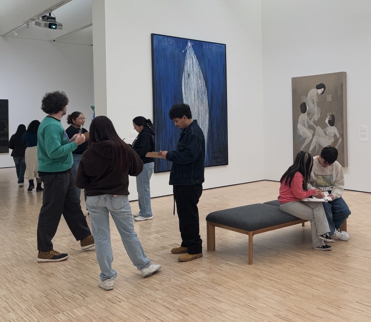 People view and discuss artwork in a modern art gallery. Some stand and converse, while two girls sit on a bench sketching or writing. Large paintings hang on white walls in a spacious, well-lit room with a wooden floor.