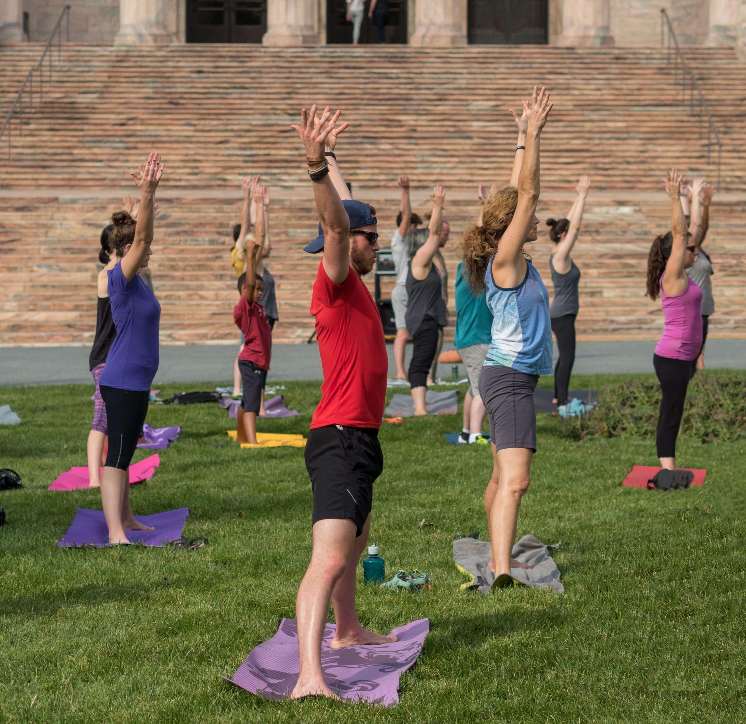 A group of people practice yoga outdoors on grass, standing on mats with arms raised overhead. They are dressed in athletic wear, and stone steps are visible in the background.