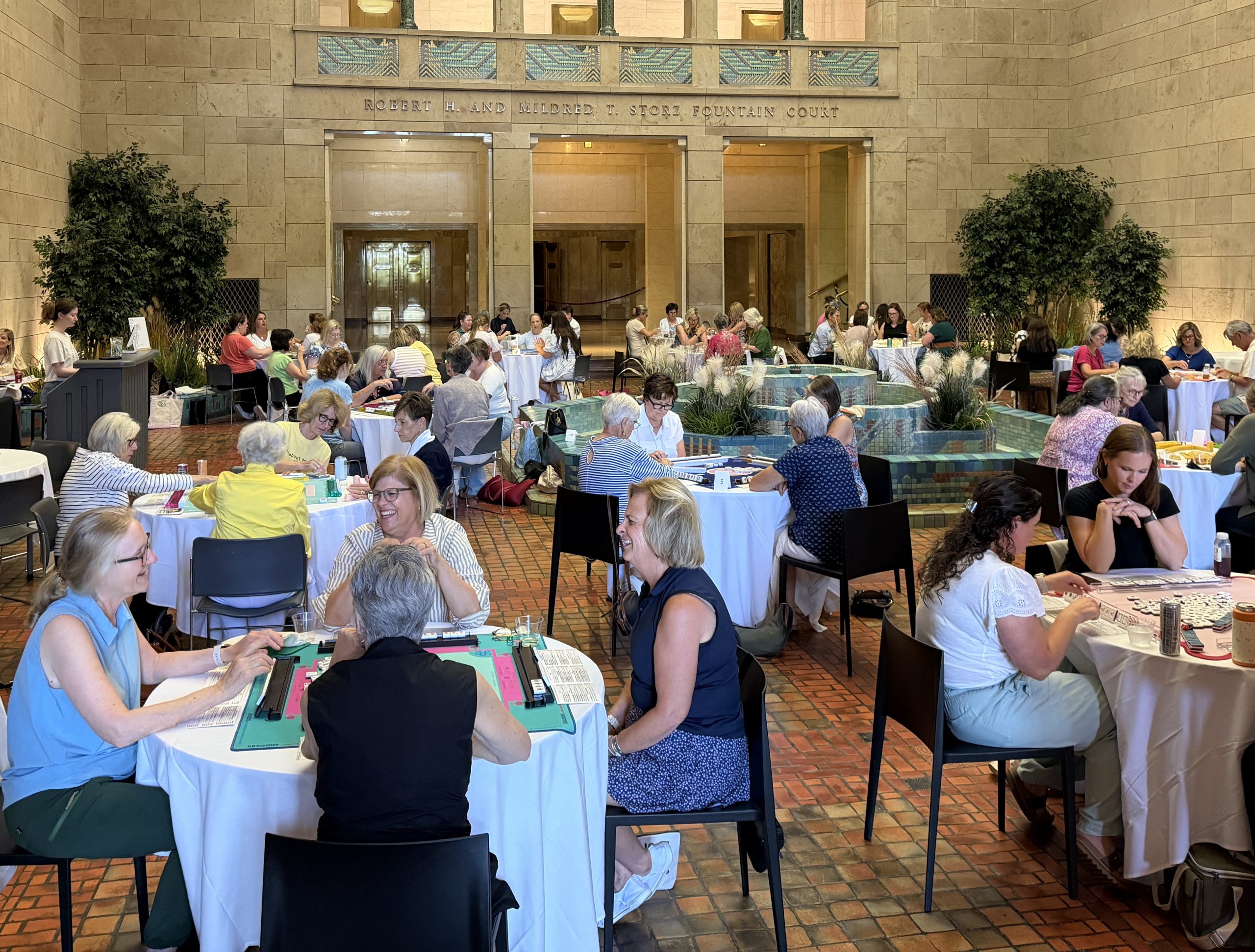 A large group of people, mostly women, sit at round tables in a bright, elegant indoor space, playing games and socializing near a central water fountain. The room has high ceilings and potted plants.