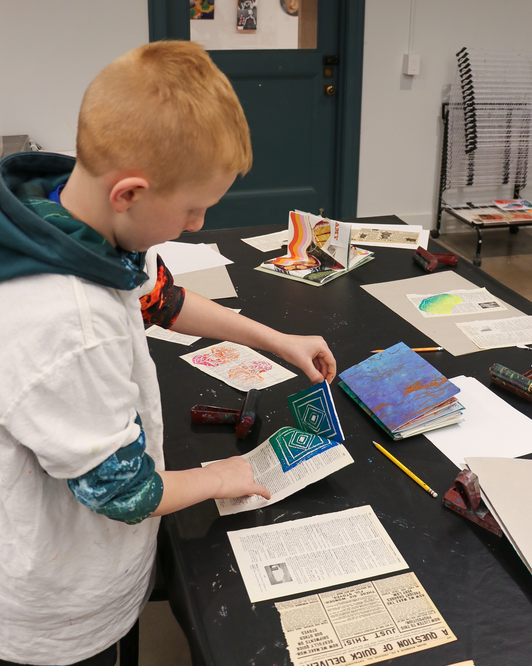 A child stands at a table, arranging colorful printed shapes and drawings on top of book pages and newspapers in an art classroom. Various art supplies and finished projects are scattered on the table.