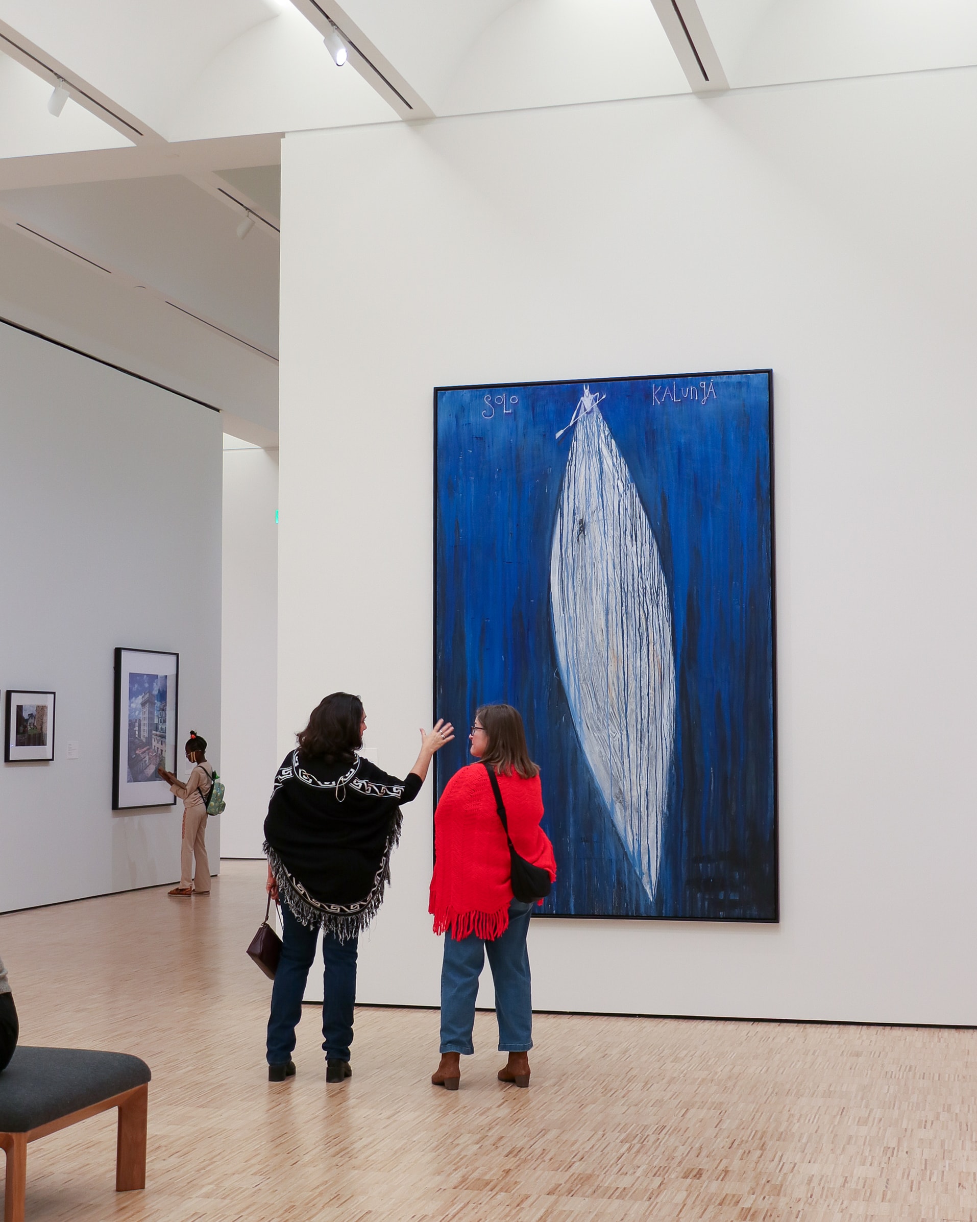 A person stands alone in a modern, minimal art gallery, looking at a large blue painting on a white wall. Sunlight streams in, casting shadows on the light wood floor.