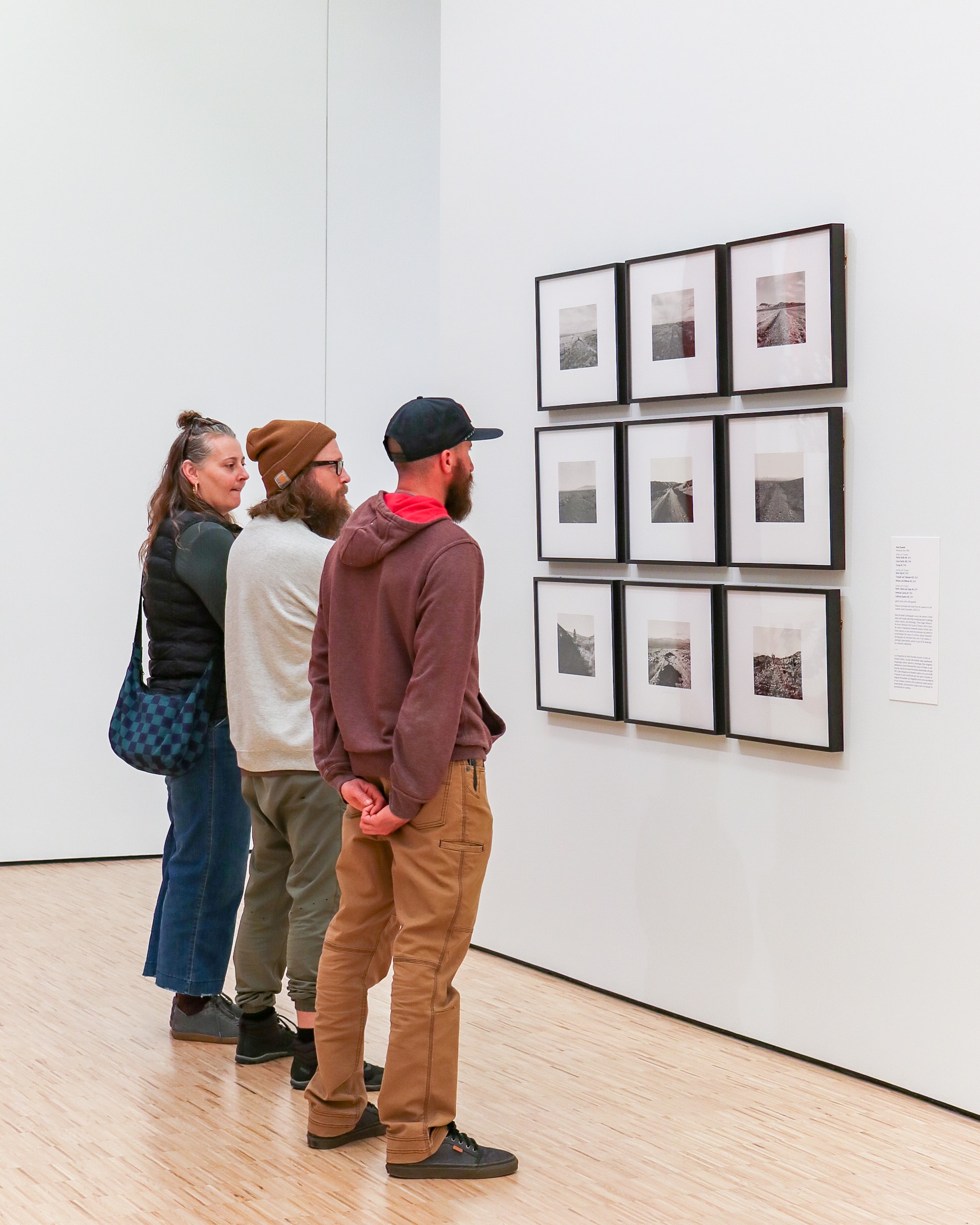 Three people stand closely together in an art gallery, looking at a wall display of nine framed black-and-white photographs arranged in a grid. The gallery has white walls and a wooden floor.