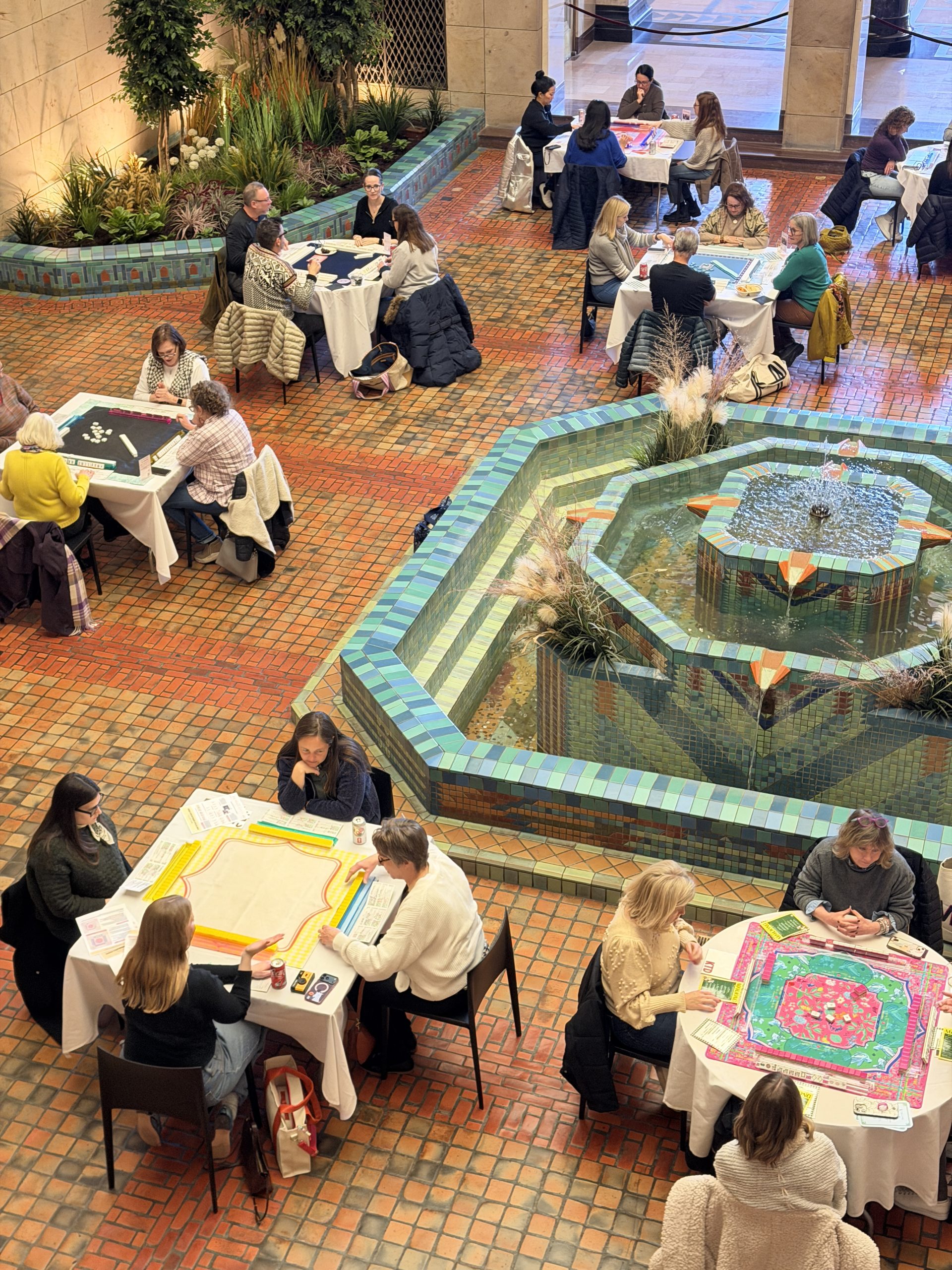 Groups of people sit at tables playing board games around a colorful tiled fountain in a spacious indoor area with brick flooring and plants. The atmosphere appears lively and social.