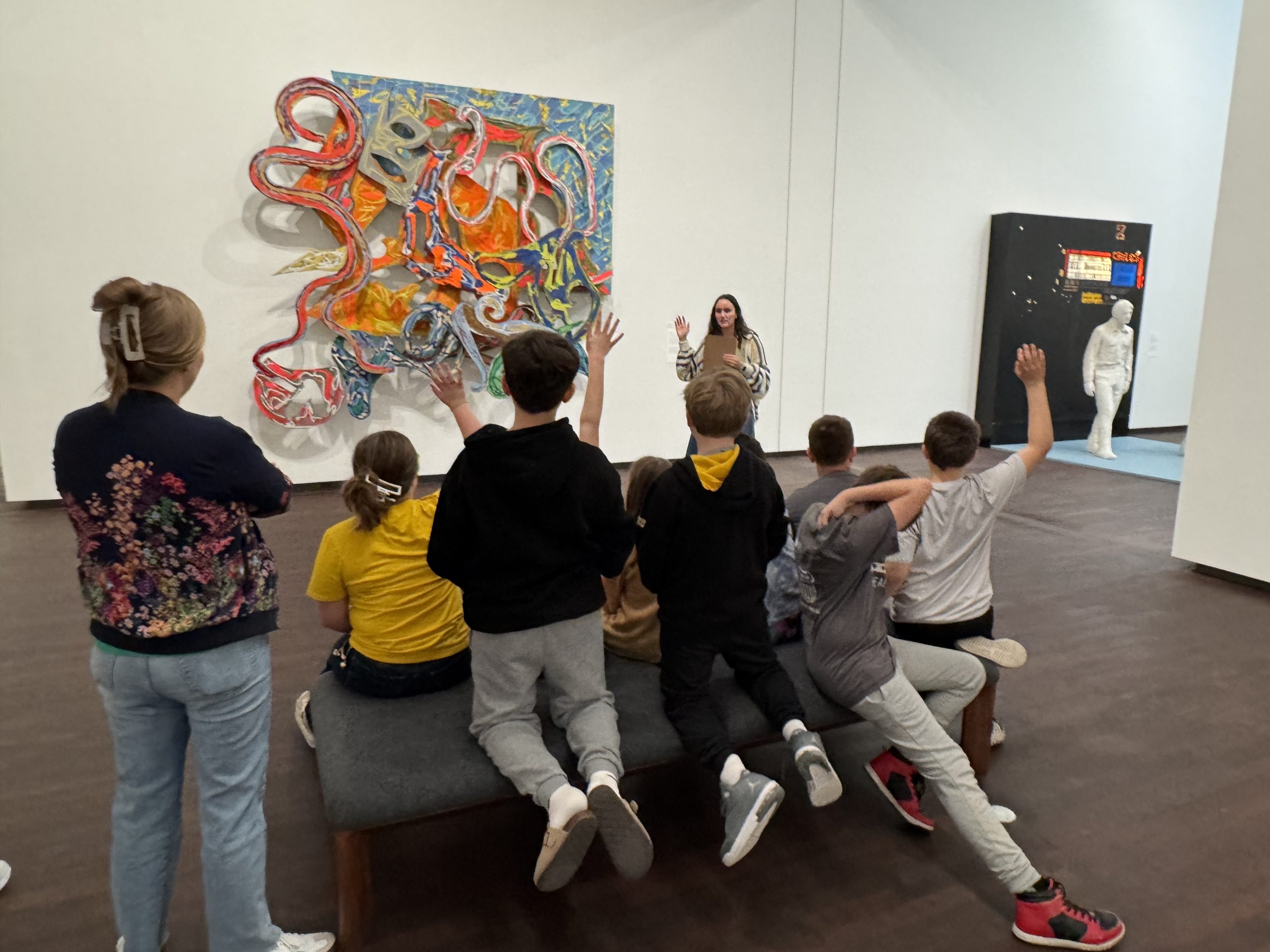 A group of children sits on a bench and raises their hands while a woman speaks near a colorful abstract wall sculpture in an art gallery. An adult stands nearby and a white statue is visible in the background.