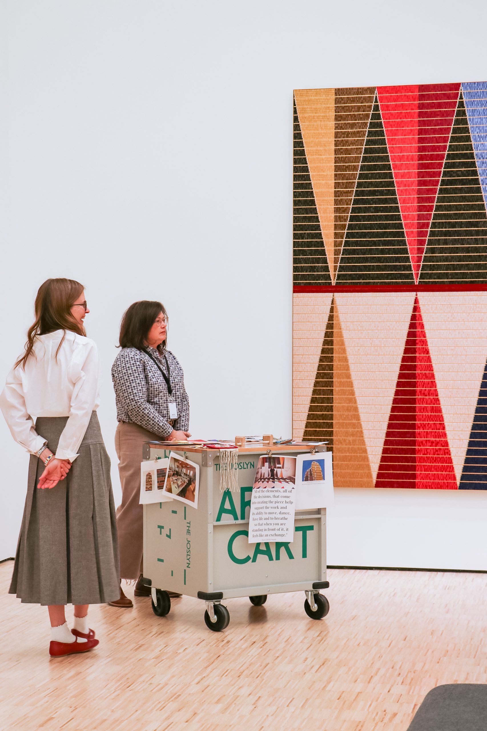 Two women stand near an art cart in a gallery, looking at a large geometric artwork with red, black, beige, and gold triangles on the wall. The cart holds art materials and displays informational signs.