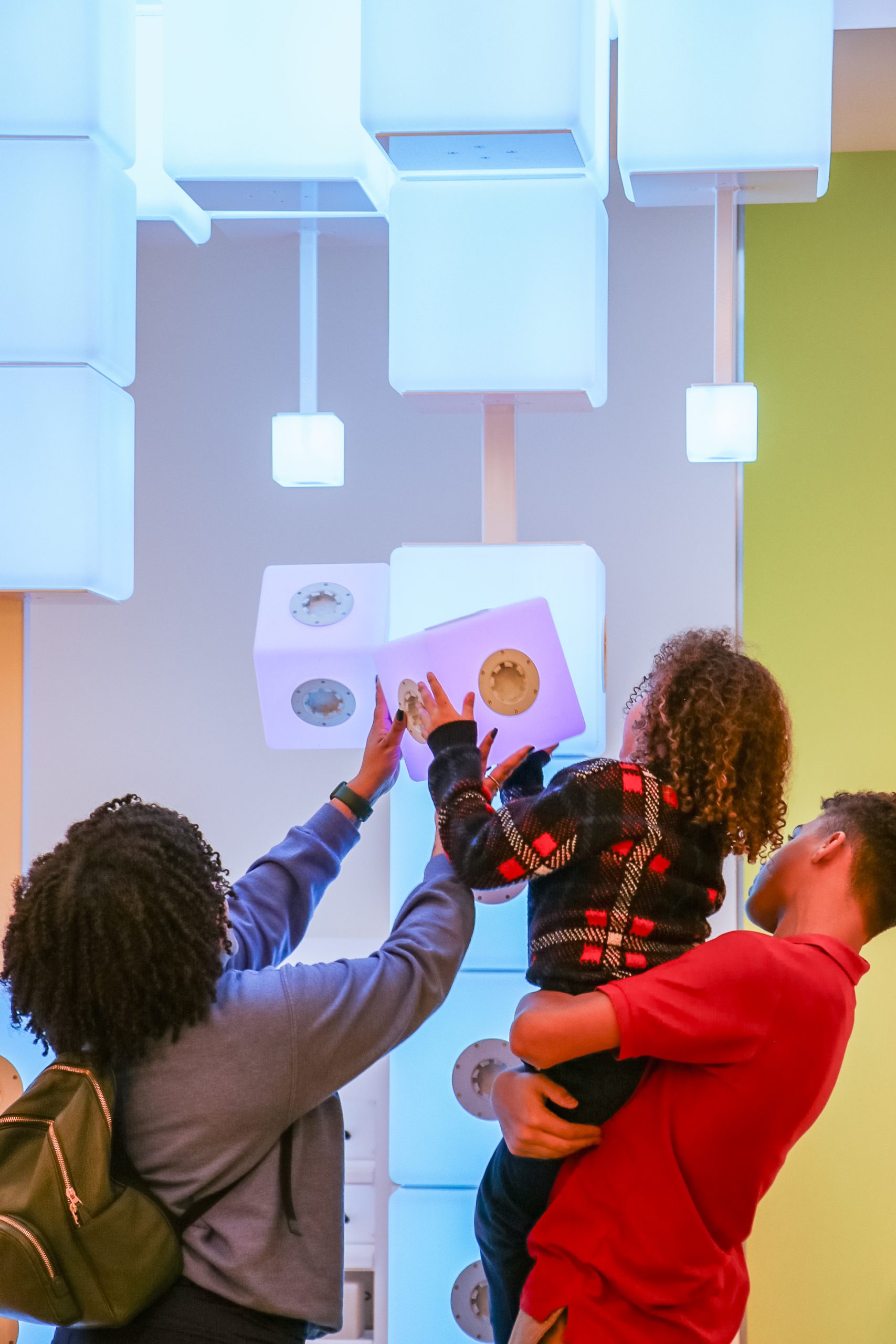Three people, including a child being held, interact with glowing, cube-shaped blocks attached to a wall in a brightly lit, modern indoor space. The scene suggests curiosity and engagement.
