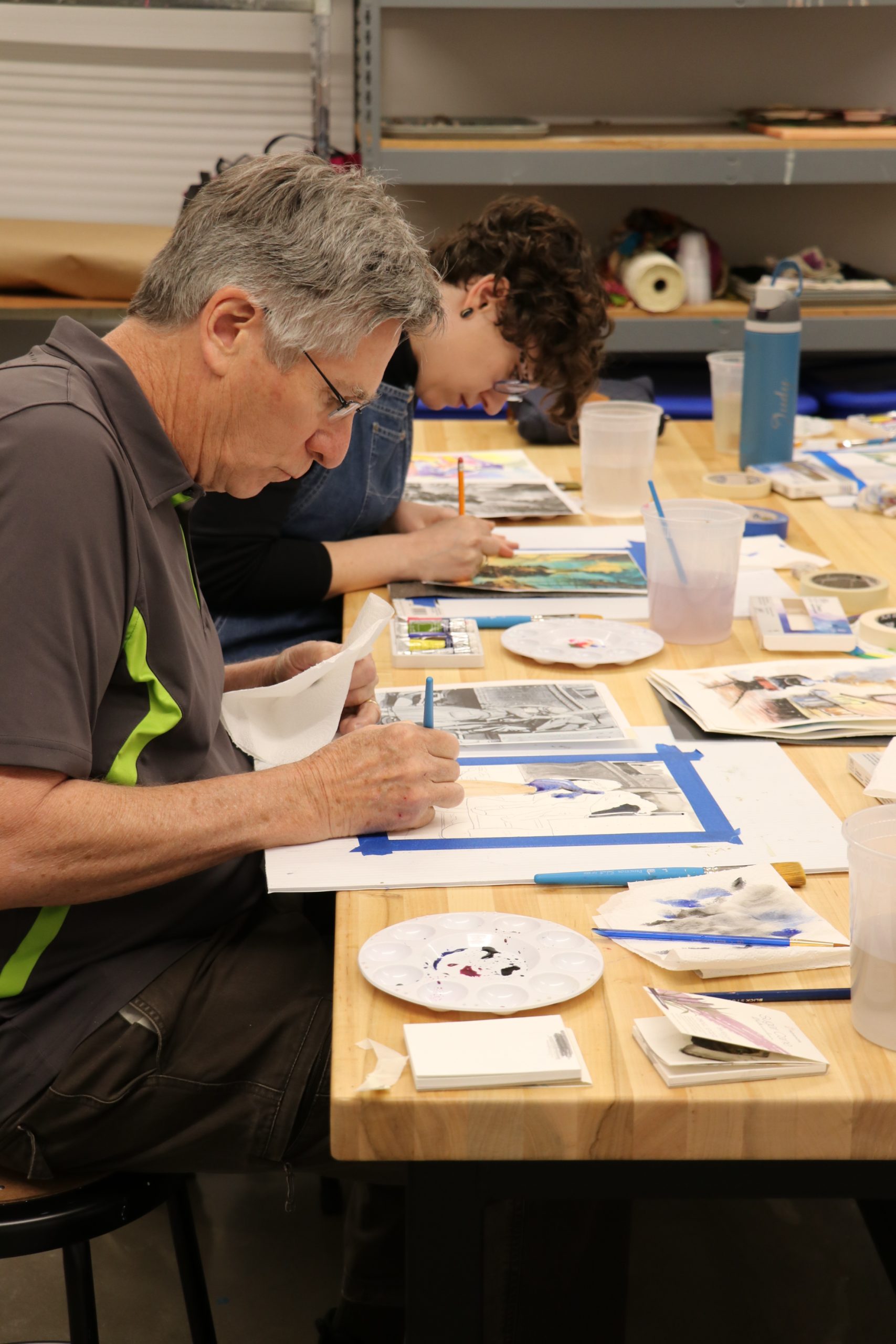 Two people sit at a wooden table, focused on painting and drawing. Art supplies, water cups, brushes, and paper are spread out on the table in a bright, organized studio environment.