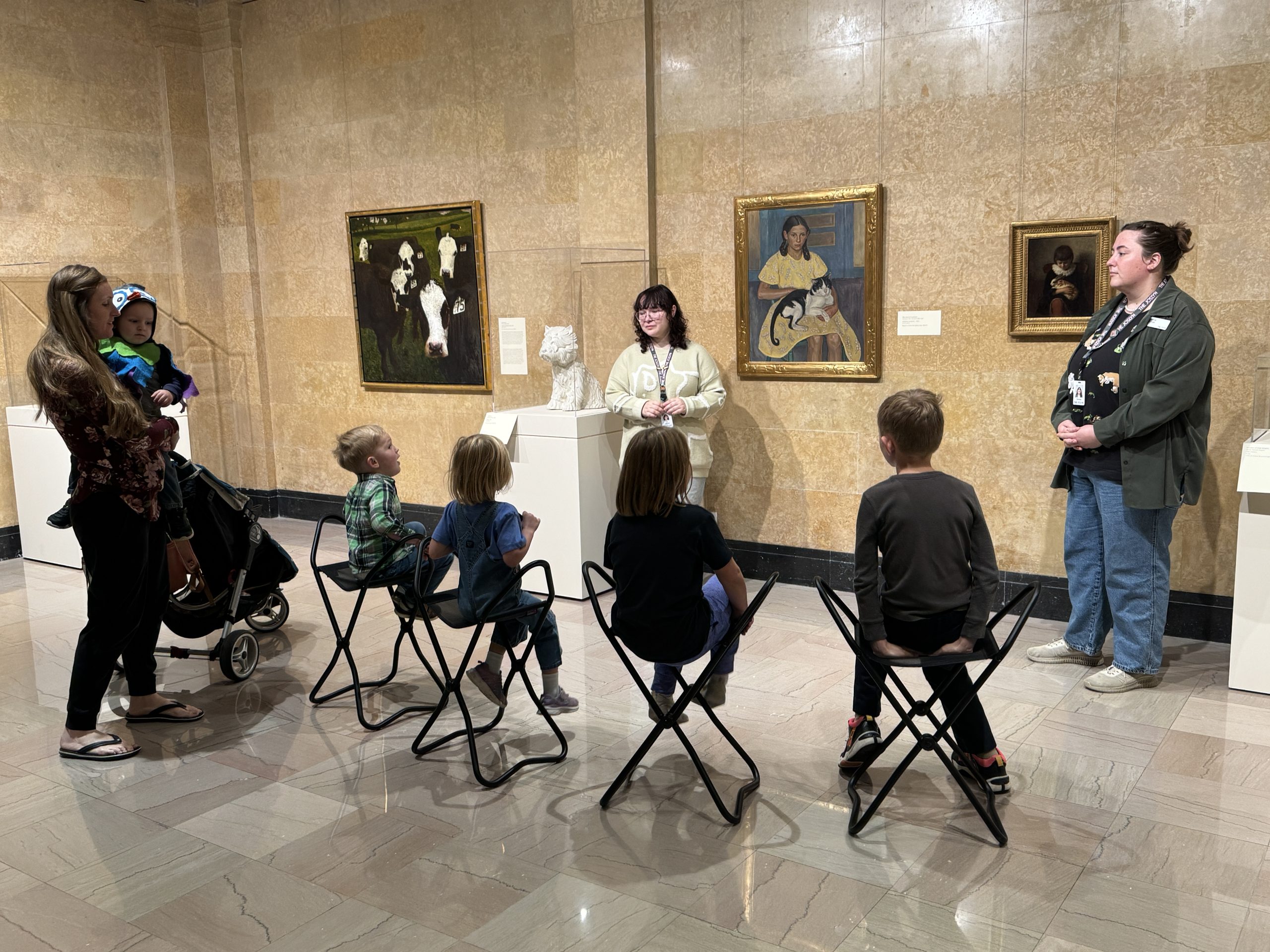 A group of children sit on folding chairs in an art museum, listening to two adult guides. A woman with a stroller and toddler stands nearby. Several paintings and a sculpture are displayed on the beige walls.