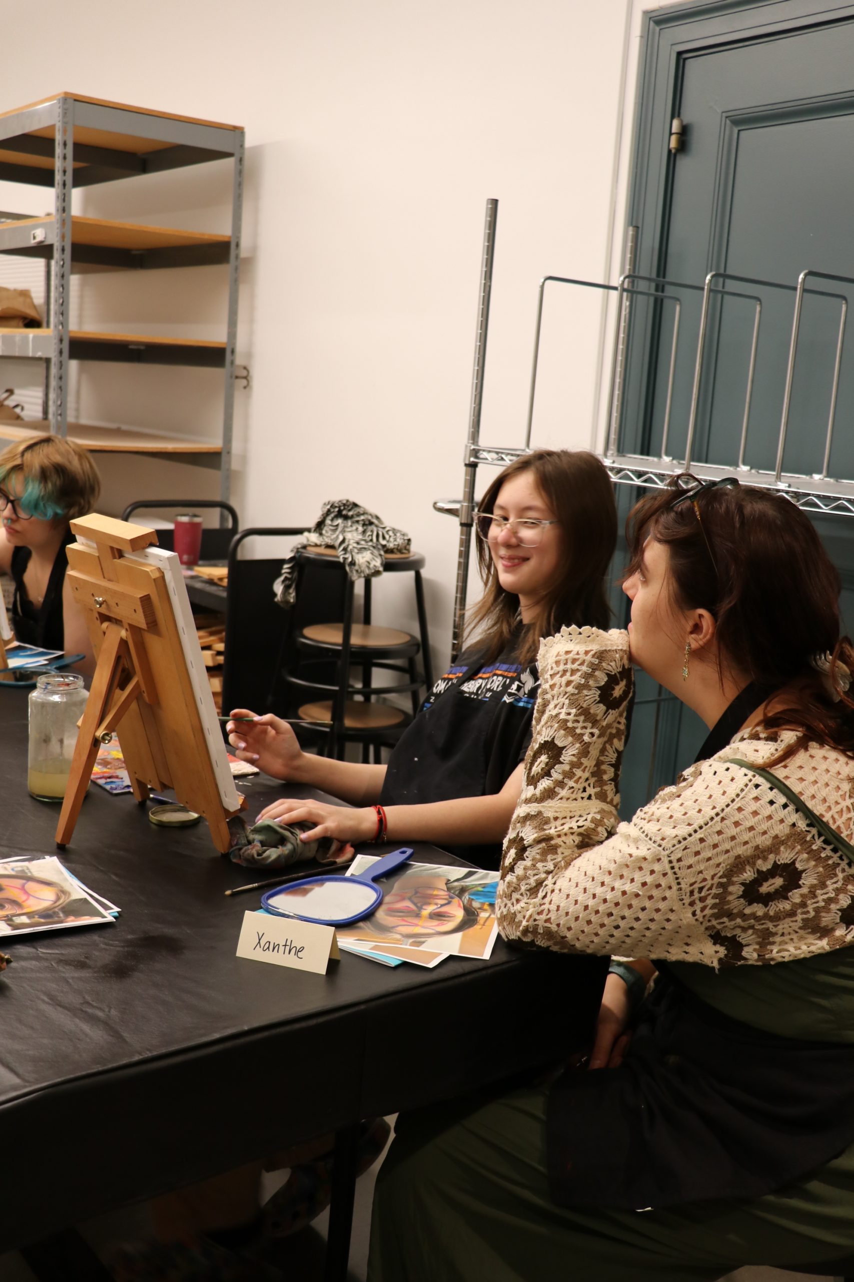 Two young women at a table with art supplies and easels. The one on the left has bright blue hair and is looking at an easel. The one on the right is smiling and wearing a patterned sweater.