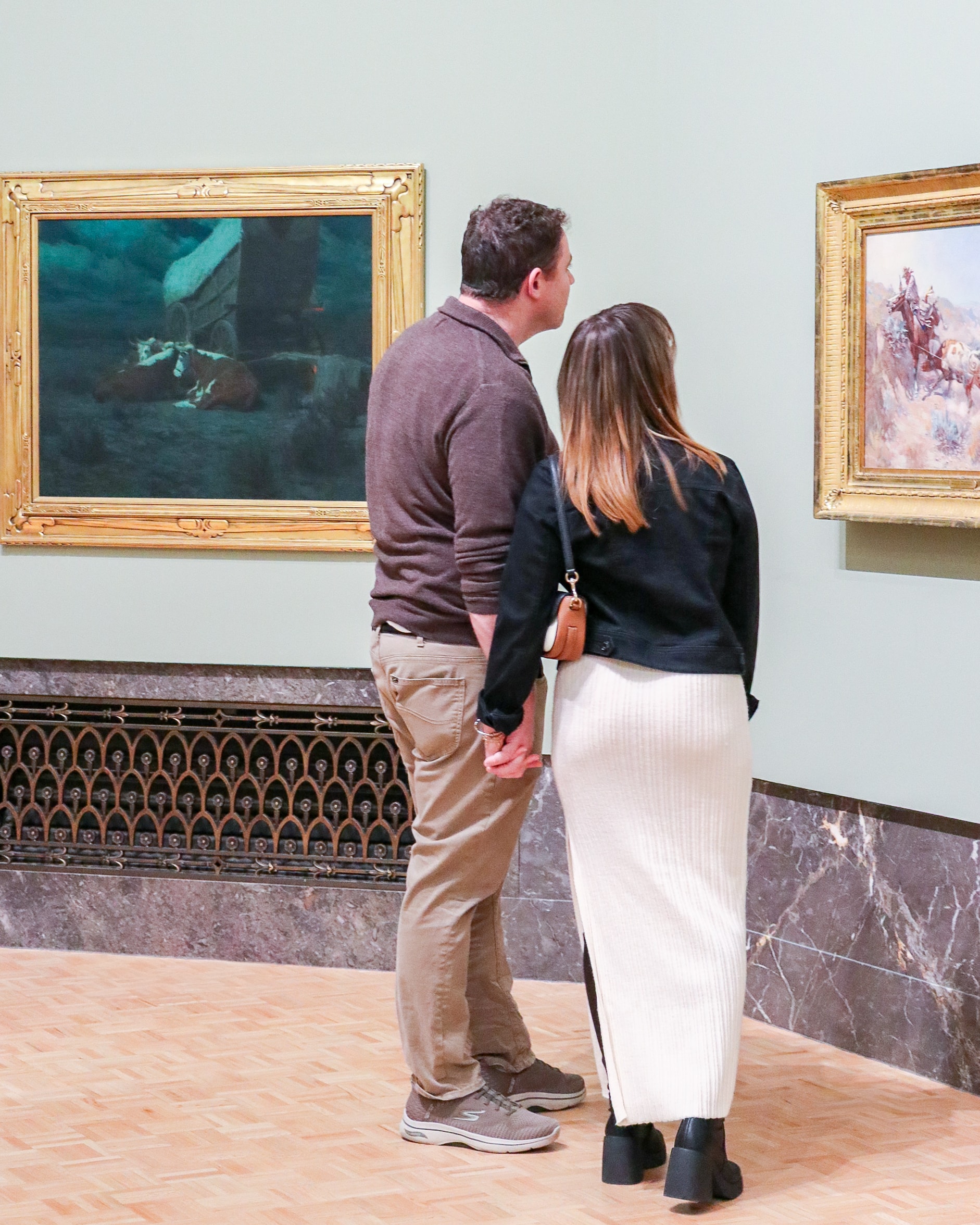 A man and woman hold hands while standing in an art gallery, viewing framed paintings on a pale green wall. The man wears a brown sweater and khaki pants; the woman wears a white skirt and black jacket.