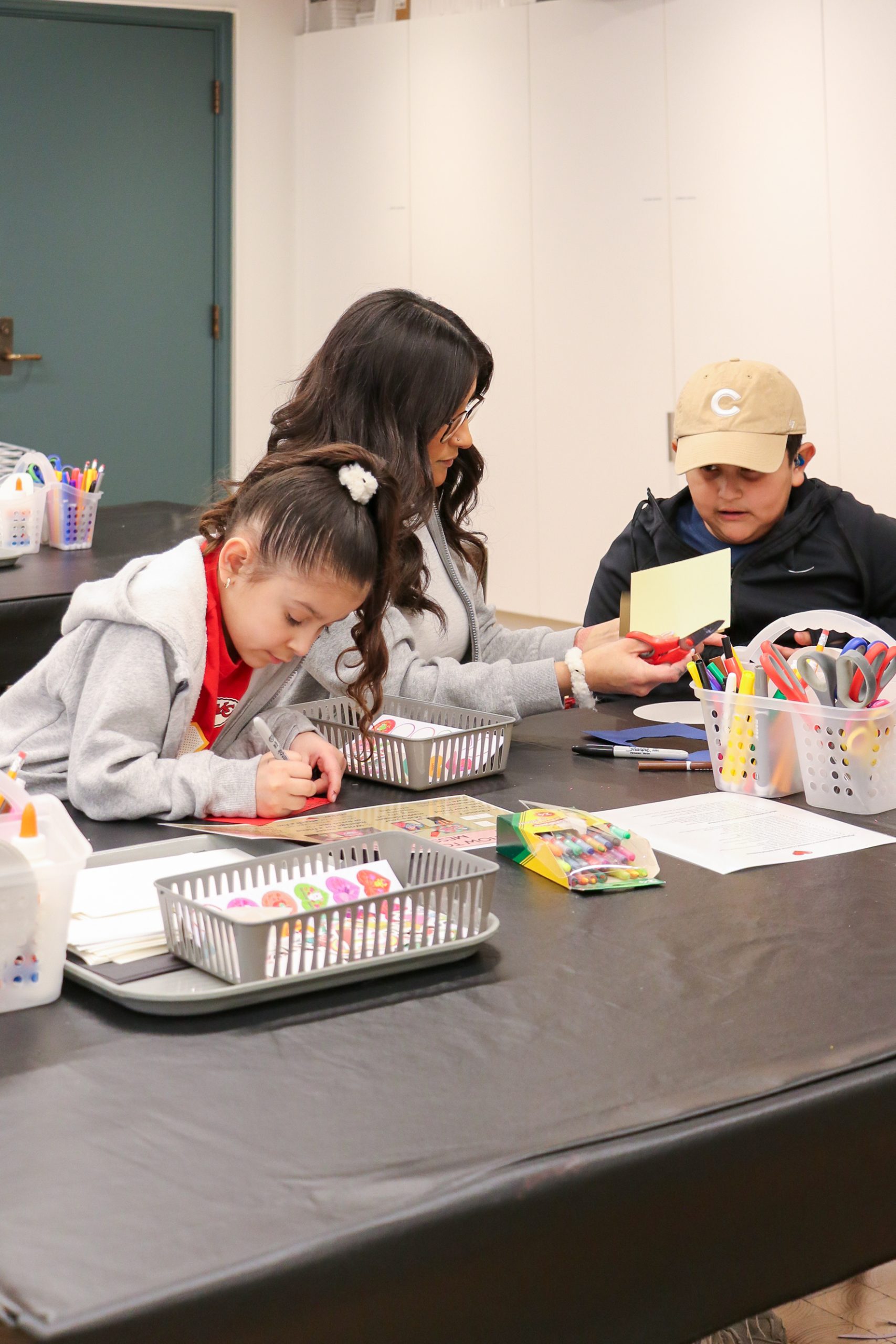 A woman and two children sit at a table doing crafts with colored markers and art supplies. The girl is drawing, while the boy in a beige cap looks at the woman, who is helping with the activity. Art materials are spread on the table.