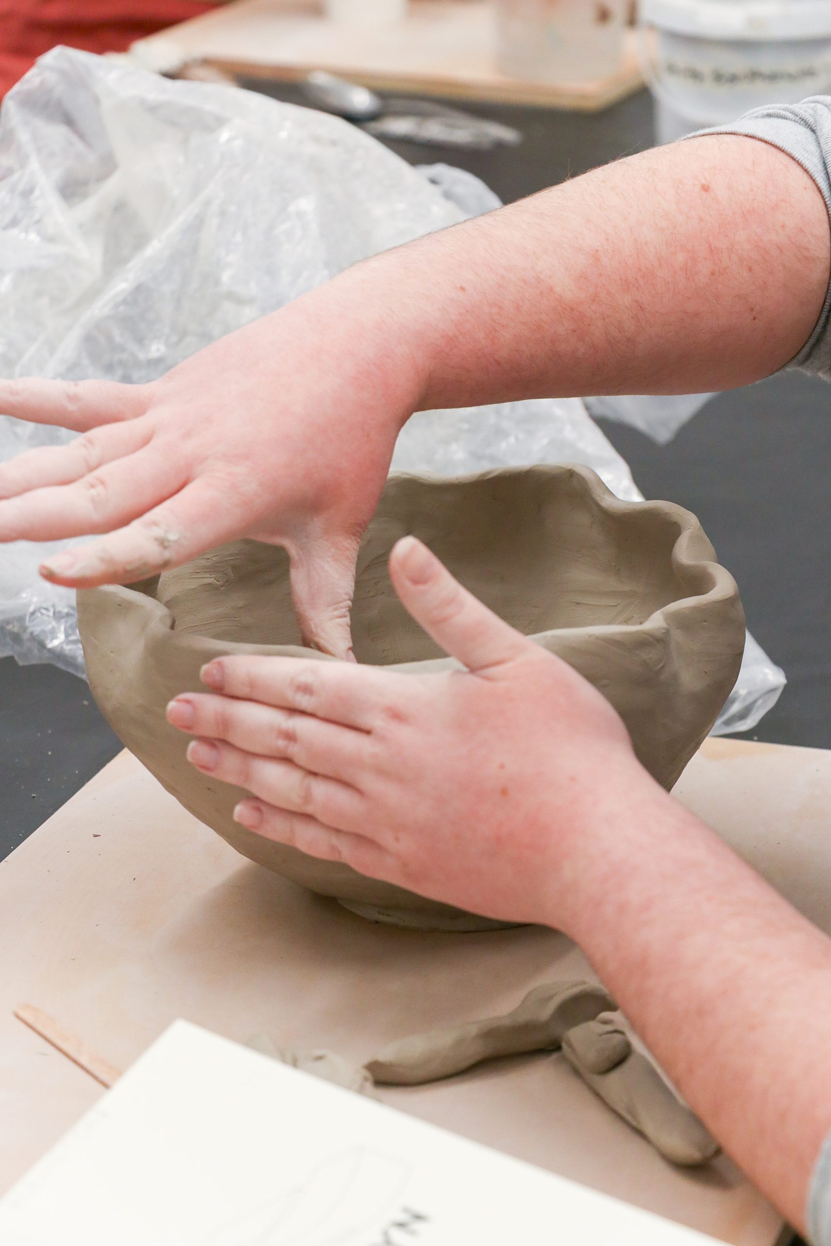 A person shapes a clay bowl with both hands on a table, surrounded by art supplies and plastic sheeting in the background.