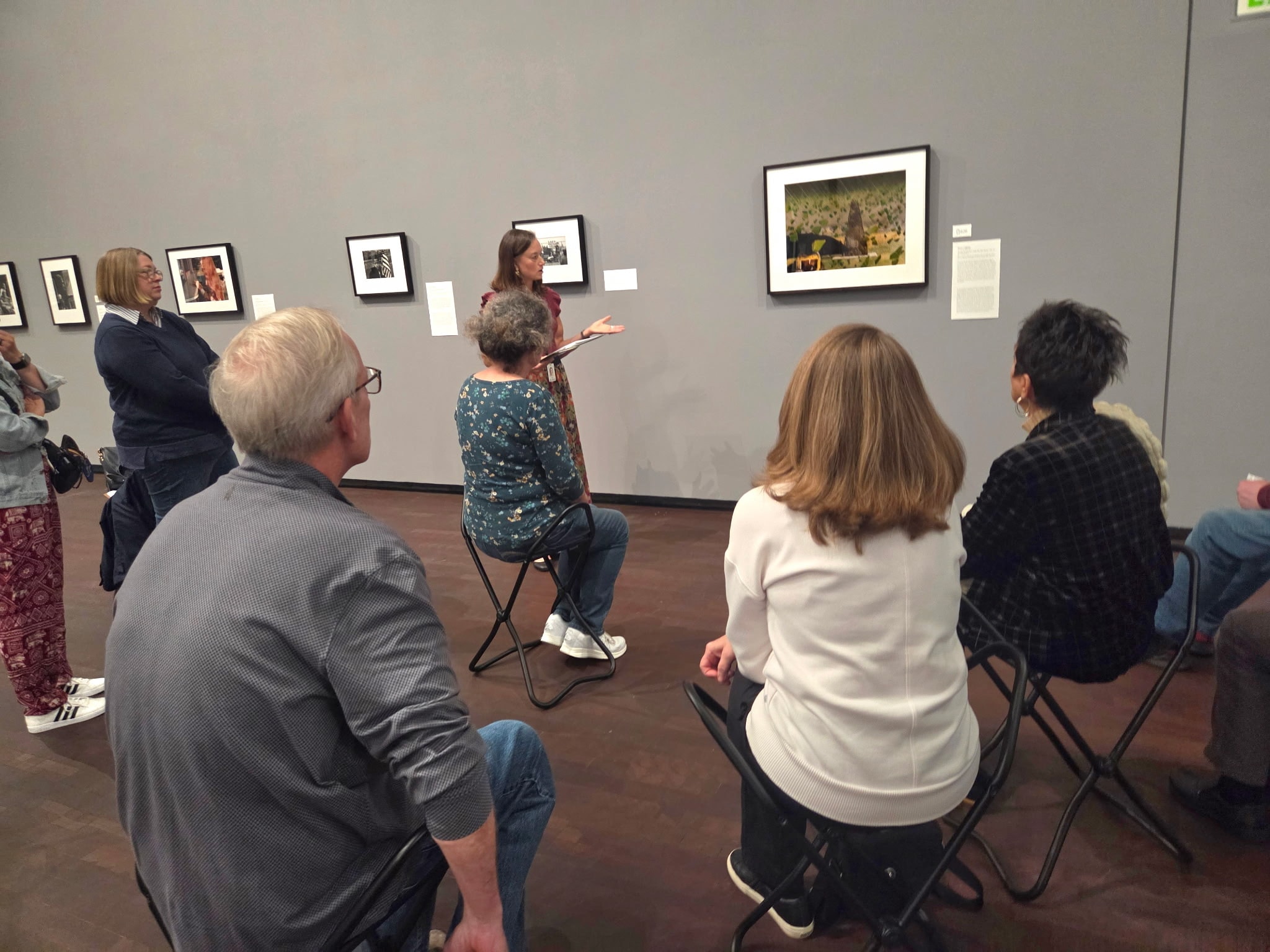A group of people sits on folding stools in an art gallery, attentively listening to a woman speaking in front of a framed photograph displayed on a gray wall. Other framed photos are visible in the background.