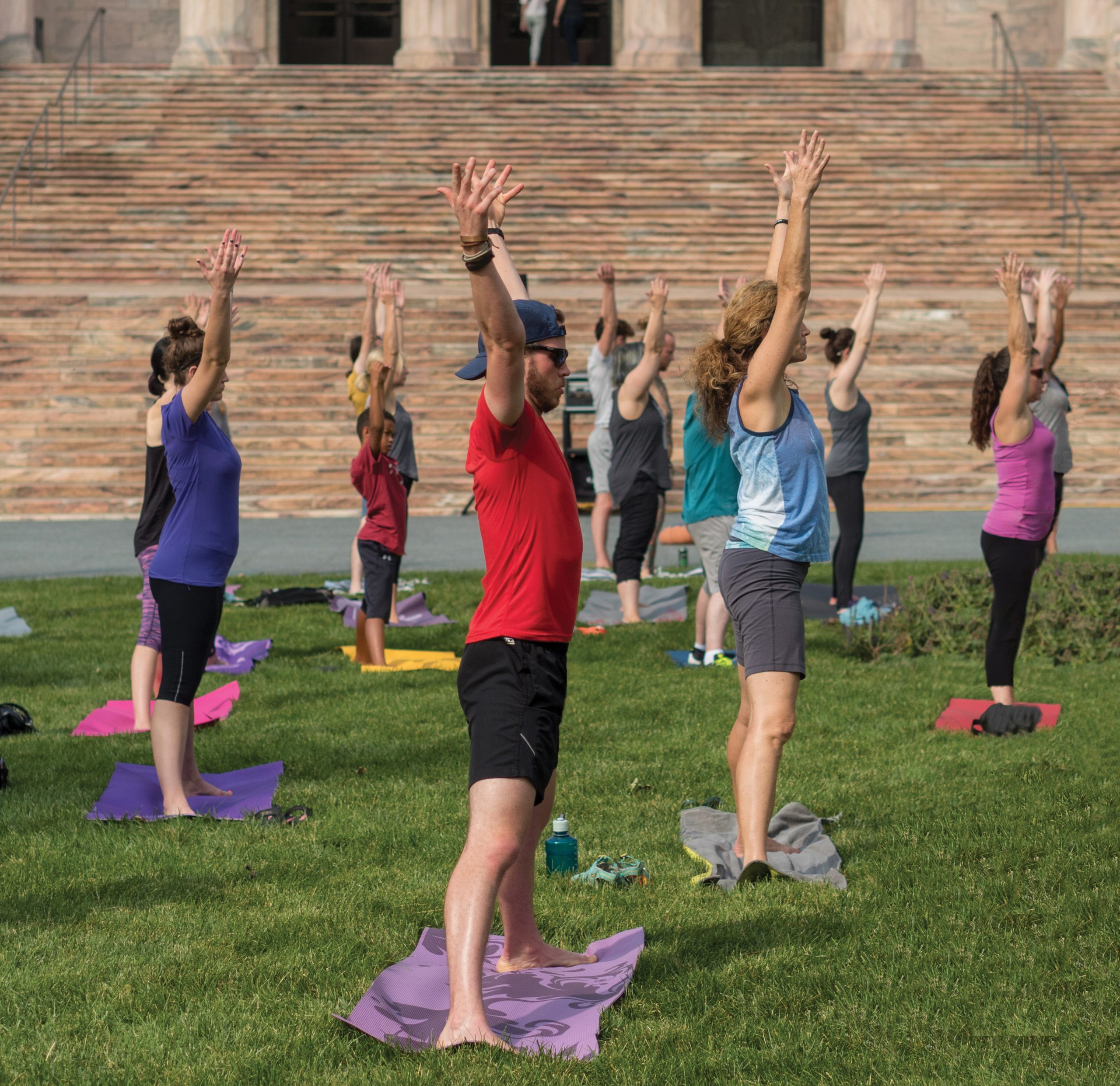 A group of people practice yoga outdoors on mats, standing with arms raised overhead on a grassy field in front of stone steps. They appear focused and aligned in their pose, enjoying an outdoor yoga class.