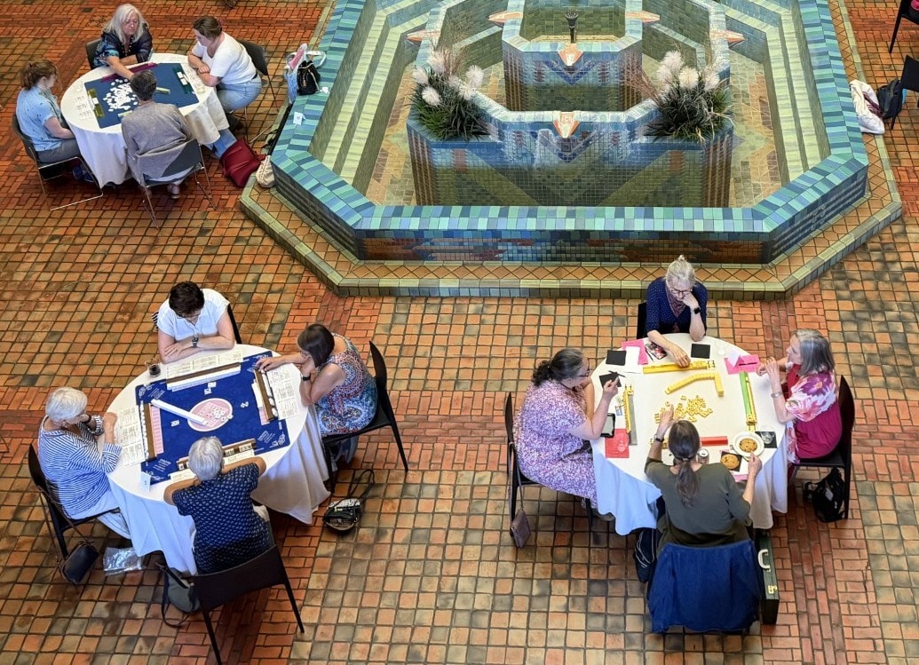 Four round tables with groups of people playing games are set around a decorative tiled fountain in a spacious, sunlit room with a brick-patterned floor.