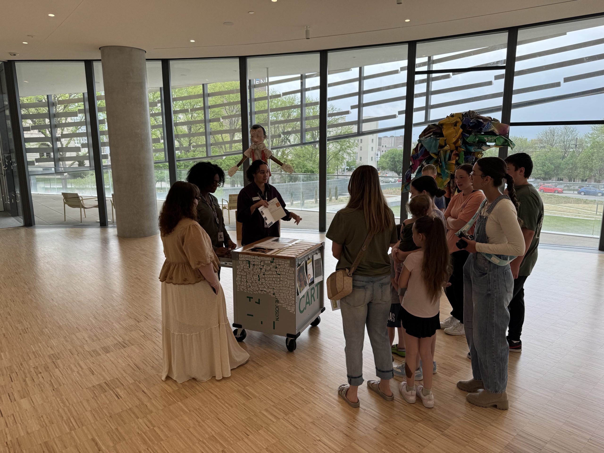 A group of people, including adults and children, gather around a cart with art supplies inside a bright, modern building with large windows and wooden floors. An instructor is speaking to the group.