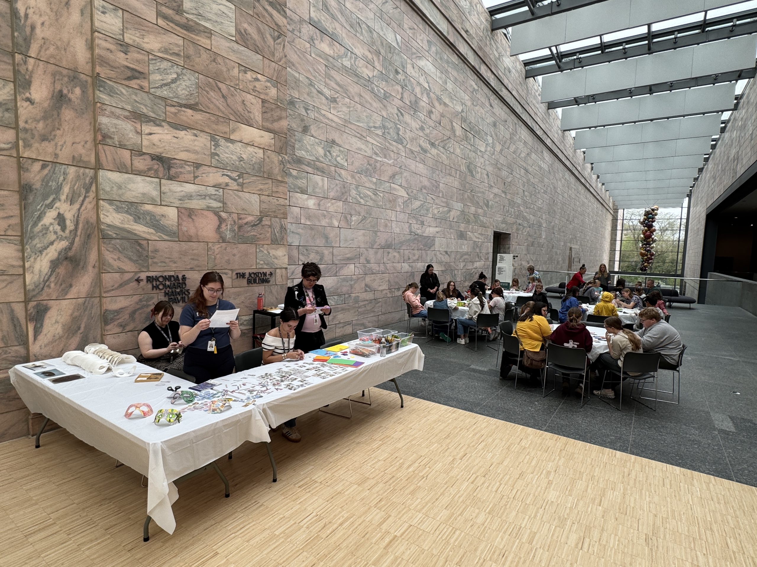 People sit at tables working on crafts in a large, modern hall with tall stone walls. Three people stand behind a table displaying art supplies and crafts. Natural light enters from the glass ceiling above.