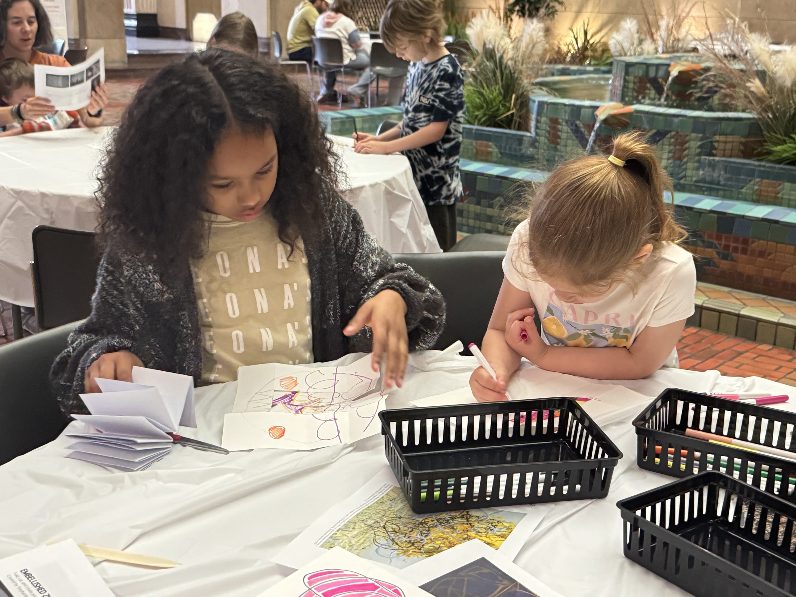 Two young girls sit at a table coloring and drawing on paper, surrounded by baskets of art supplies. Other people work at tables in the background near a tiled fountain and plants.