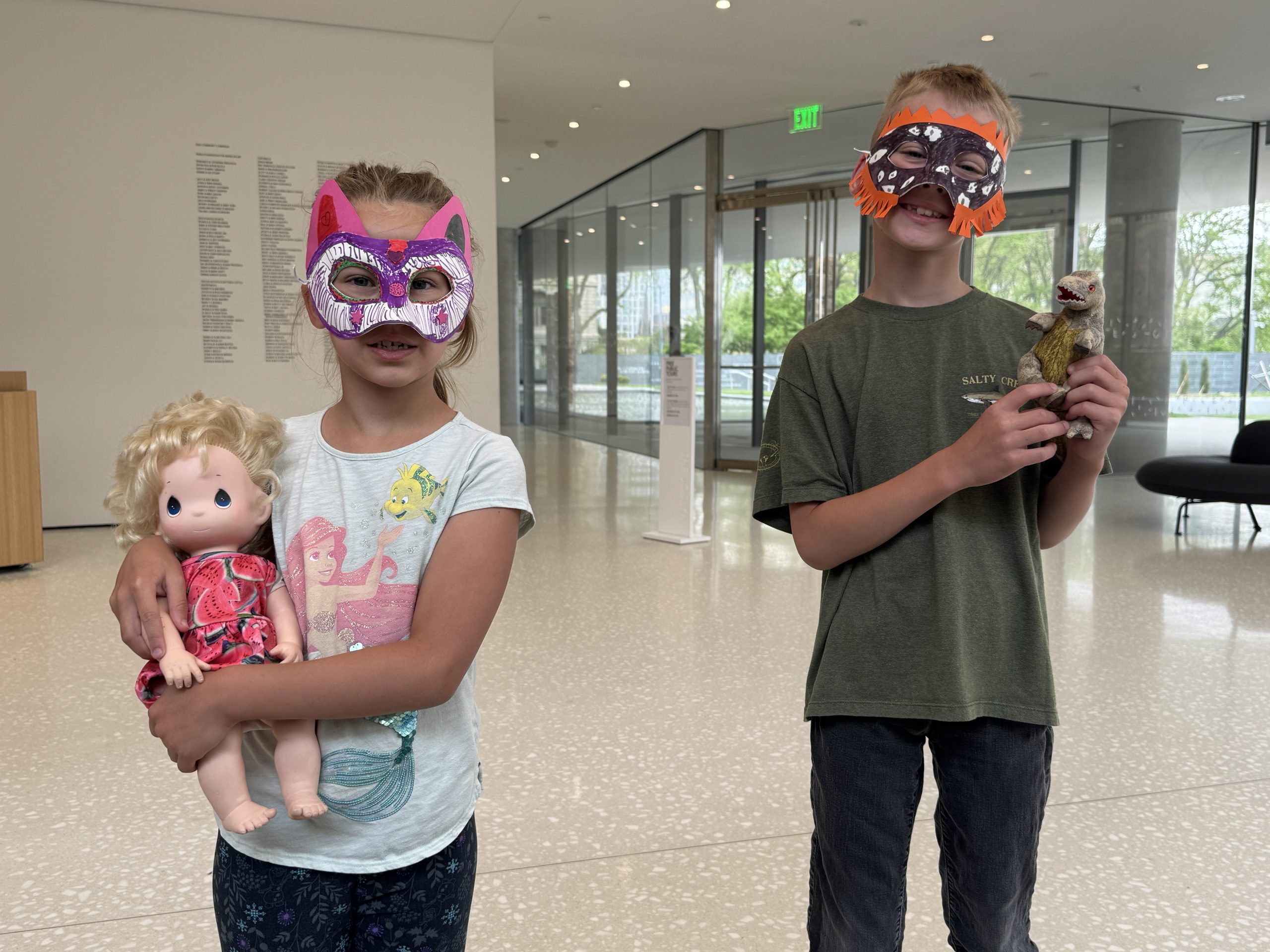 Two children stand indoors wearing colorful animal masks. The girl holds a blonde doll and wears a cat mask, while the boy holds a small animal toy and wears a brown mask. The room has large windows and modern decor.