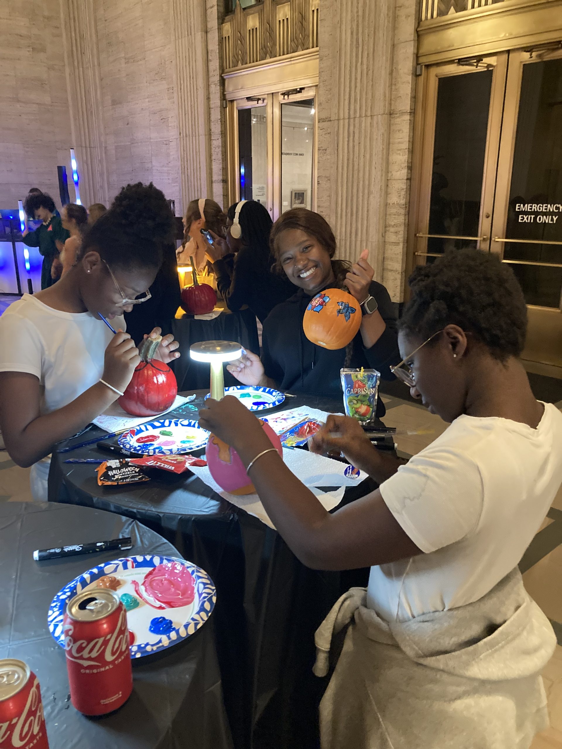 Three people sit at a table indoors, painting pumpkins and smiling. The table has art supplies, snacks, and soda cans. Warm light illuminates their workspace. Others are visible in the background.