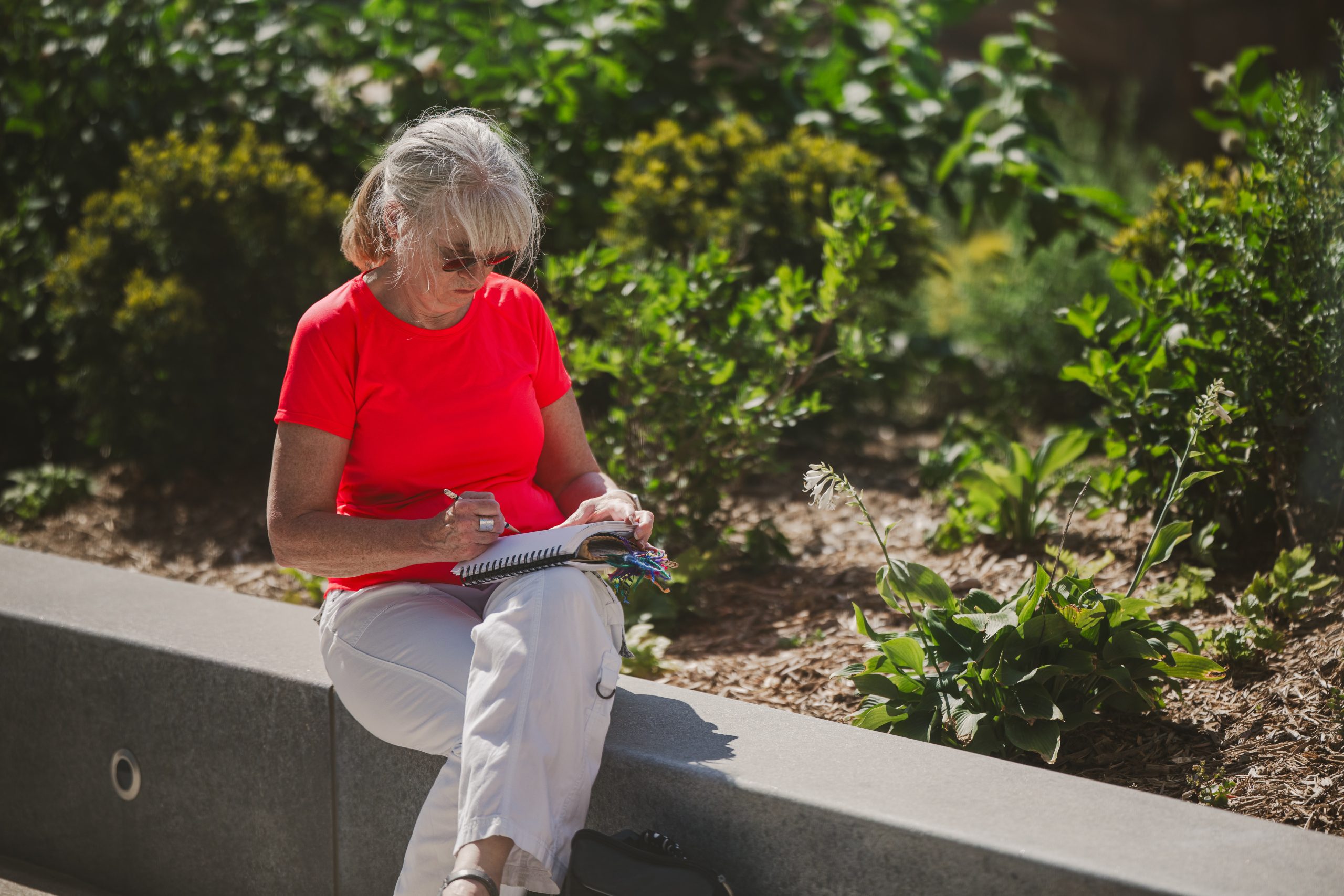 An older woman with gray hair, wearing a bright red shirt and white pants, sits on a concrete bench outside, writing in a notebook, surrounded by green plants and sunlight.