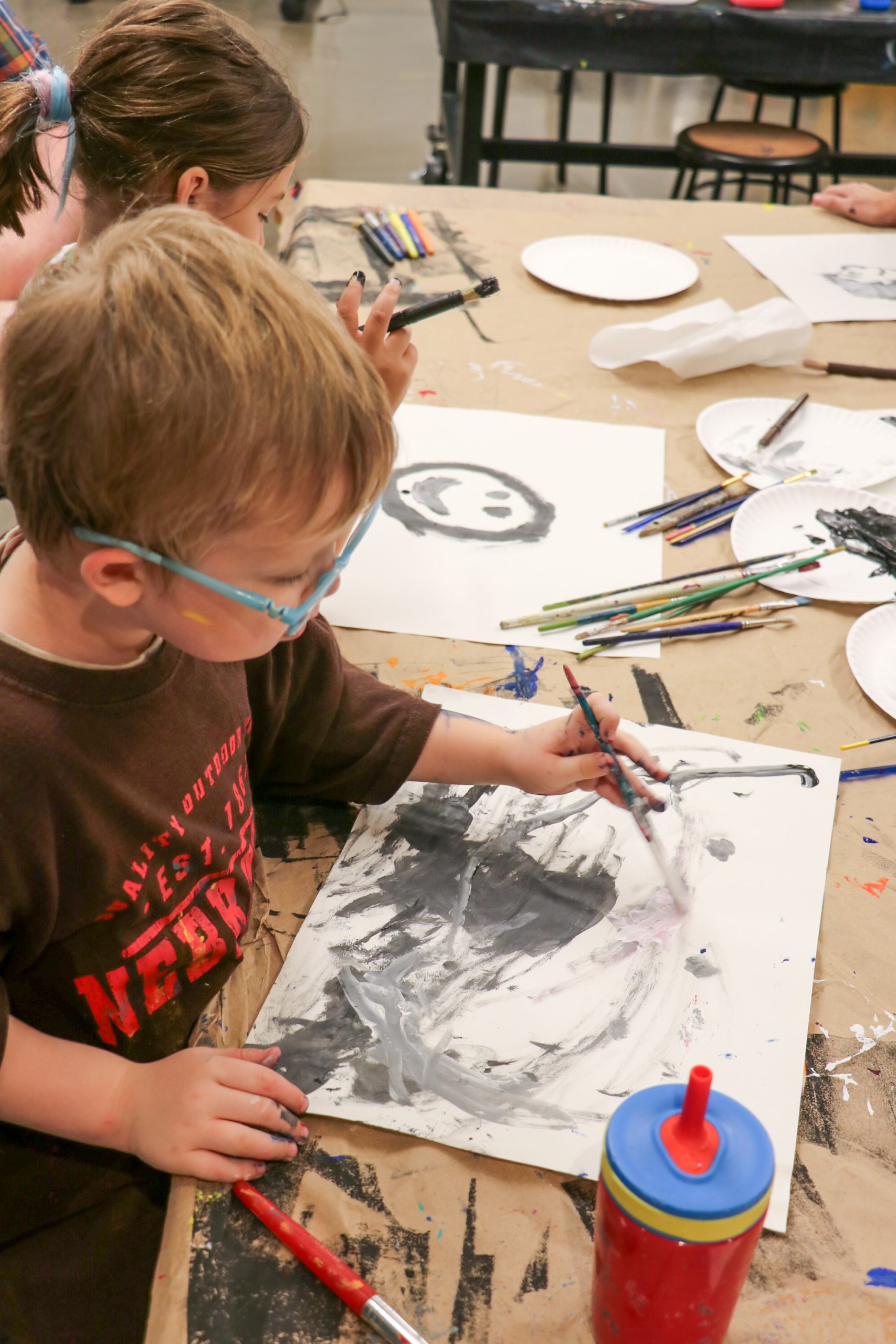 Young child with glasses painting on paper at a table with art supplies, another child painting nearby, colorful crayons scattered, and a water bottle in foreground.