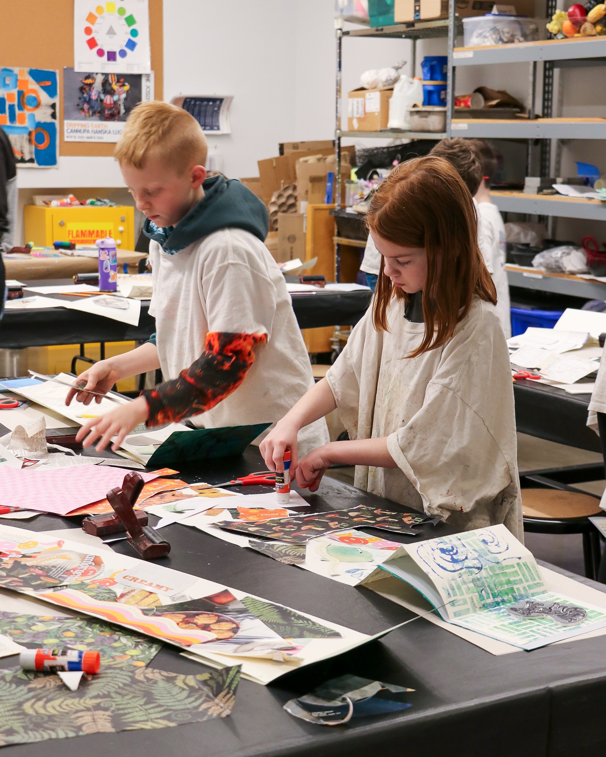 Two children in an art classroom use glue sticks to create colorful collages with paper cutouts on a table covered in art supplies. Shelves with bins and art projects are visible in the background.