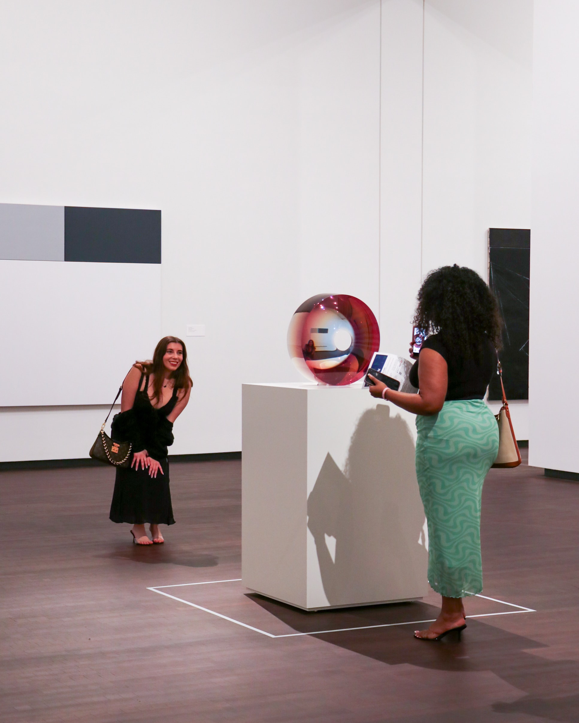 Two women interact with a spherical sculpture on a pedestal in an art gallery. One bends and smiles, while the other stands, holding a phone or brochure. Minimalist art hangs on white walls around them.