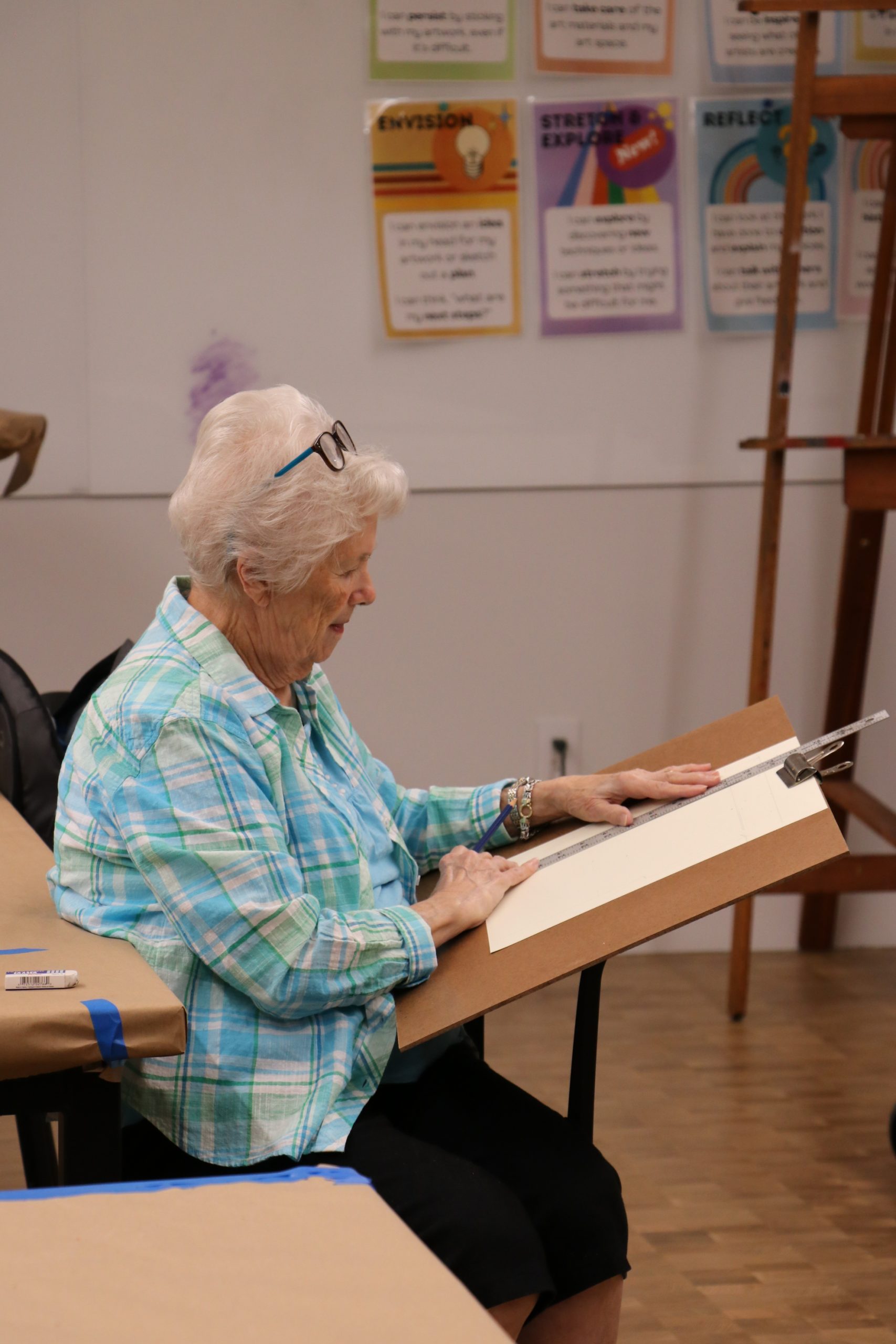 An elderly woman sits and sketches on a drawing board in an art classroom. Colorful posters with motivational phrases hang on the wall behind her.