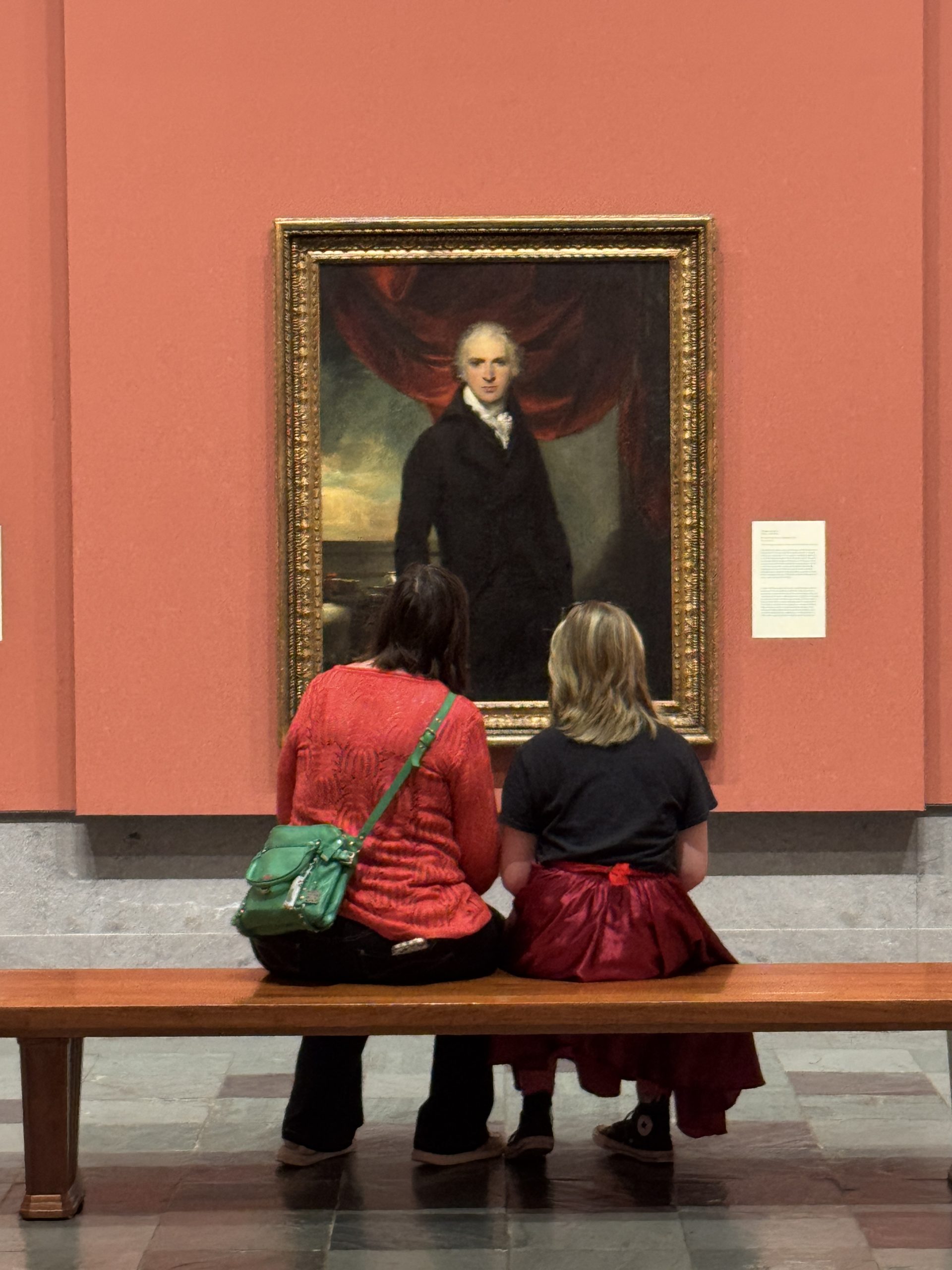 Two people sit on a bench in an art gallery, facing a large framed portrait of a man in historical attire displayed on a salmon-colored wall.
