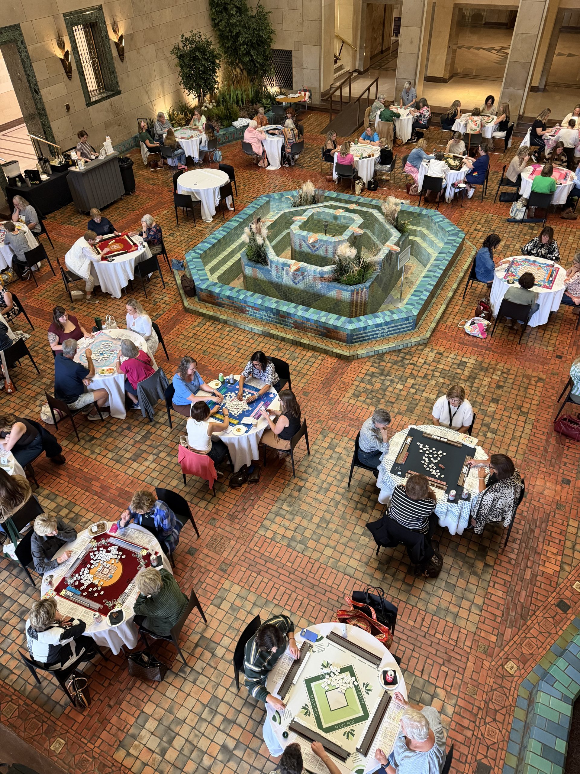 People sit in groups at round tables playing board games in a spacious indoor atrium. A decorative fountain with plants is in the center, and the floor is covered in reddish-brown tiles.