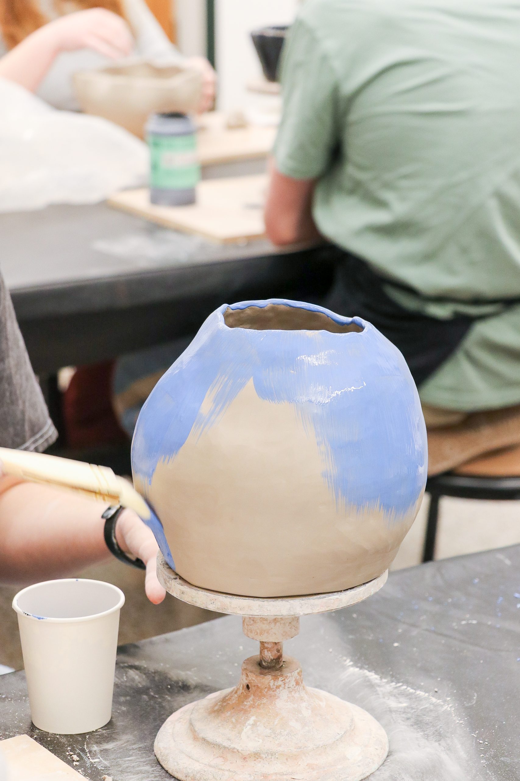 A person paints blue glaze onto a round clay pot placed on a pottery wheel. A cup and paintbrush are on the table, with other people working in the background.