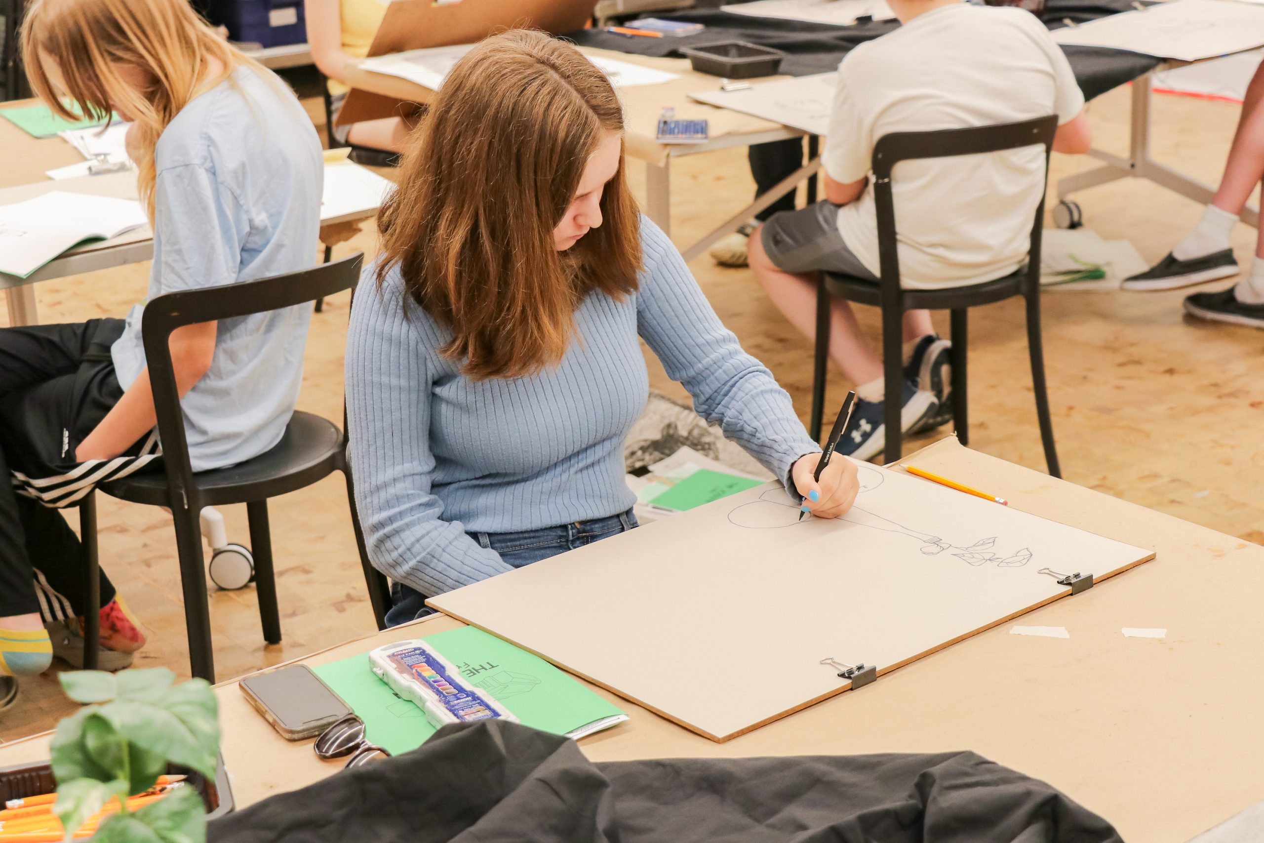 A young woman with brown hair draws on a large sketchpad at a table in an art classroom. Other students are also drawing around her, and art supplies are scattered on the tables.