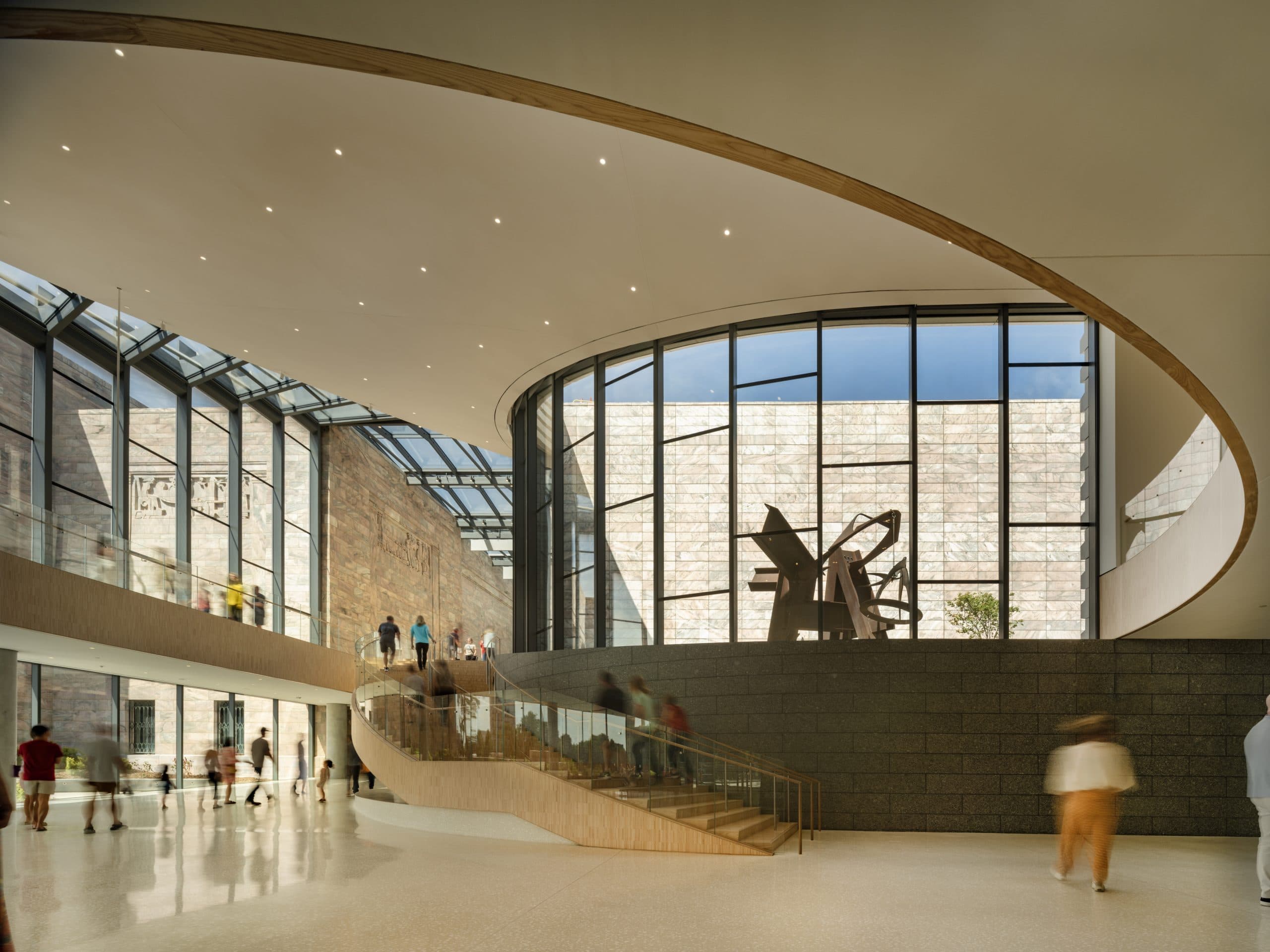Spacious modern museum interior with a large curved staircase and tall glass windows. Visitors walk around, admiring the space. A large sculpture is visible through the windows. The ceiling features recessed lighting.