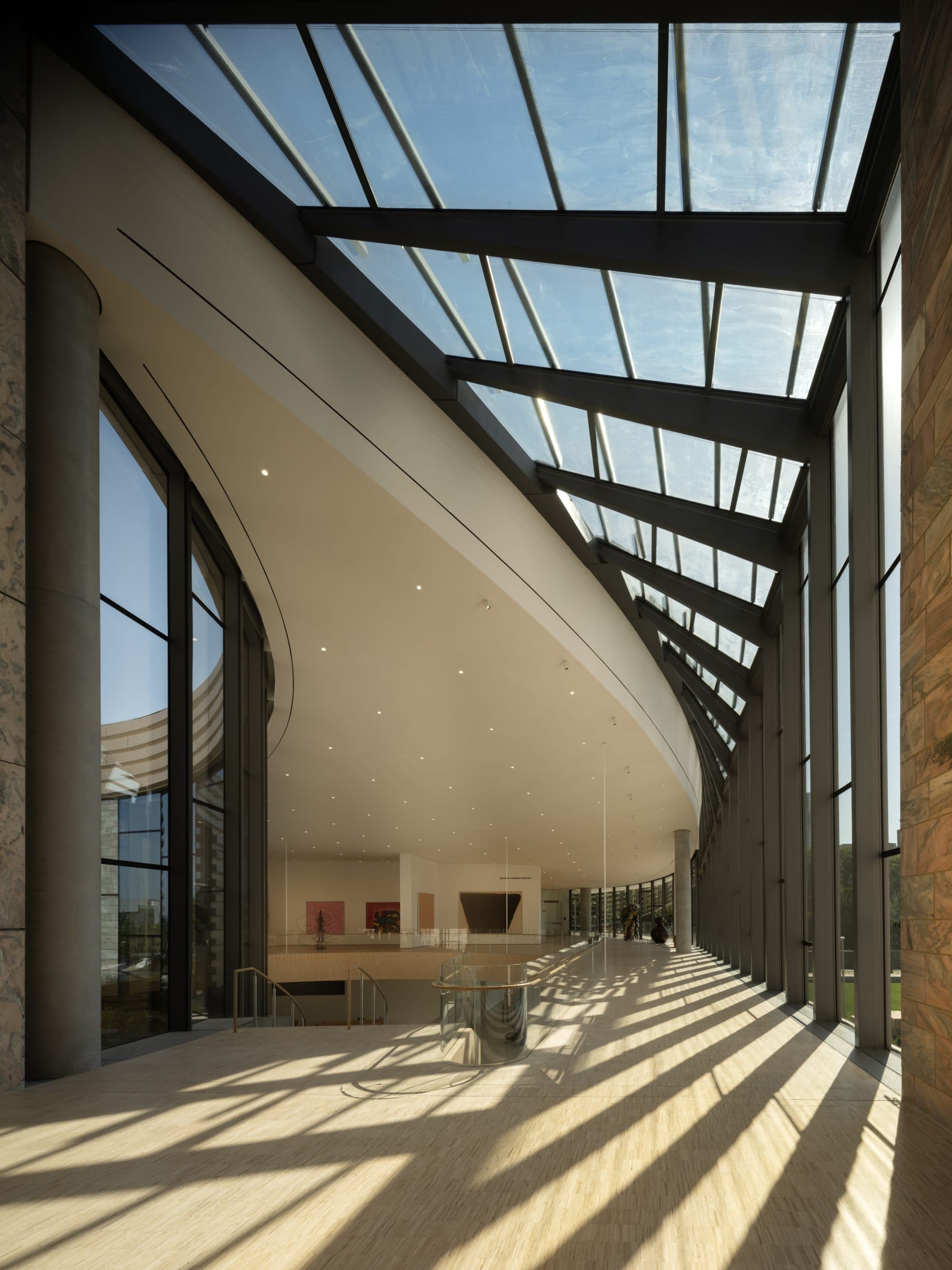 Spacious modern atrium with large floor-to-ceiling windows and a glass ceiling, allowing natural light to stream in. The interior features sleek lines, minimalist design, and light-colored flooring, creating an open and airy atmosphere.