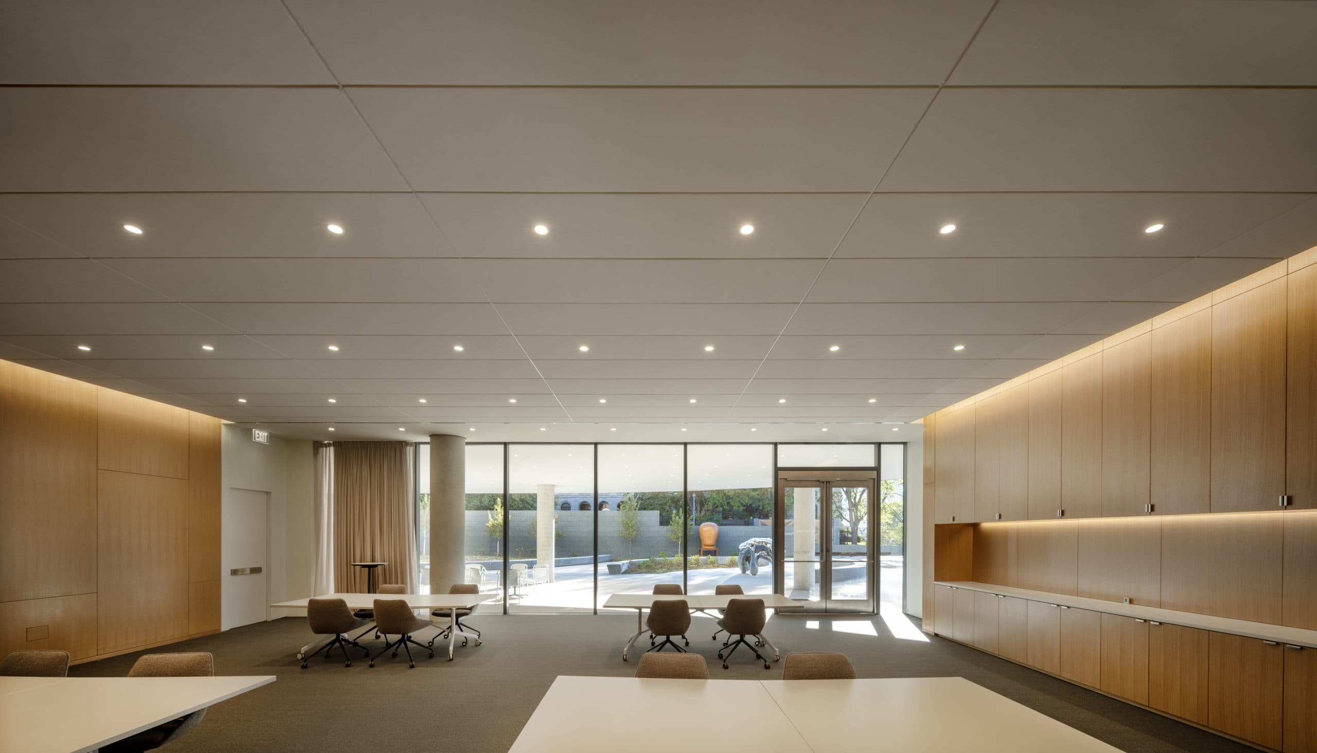 Minimalist conference room with modern ceiling lights, large windows, and natural light. Brown chairs and tables are arranged around the room. The back wall features wooden paneling with built-in storage. Outside, greenery is visible.