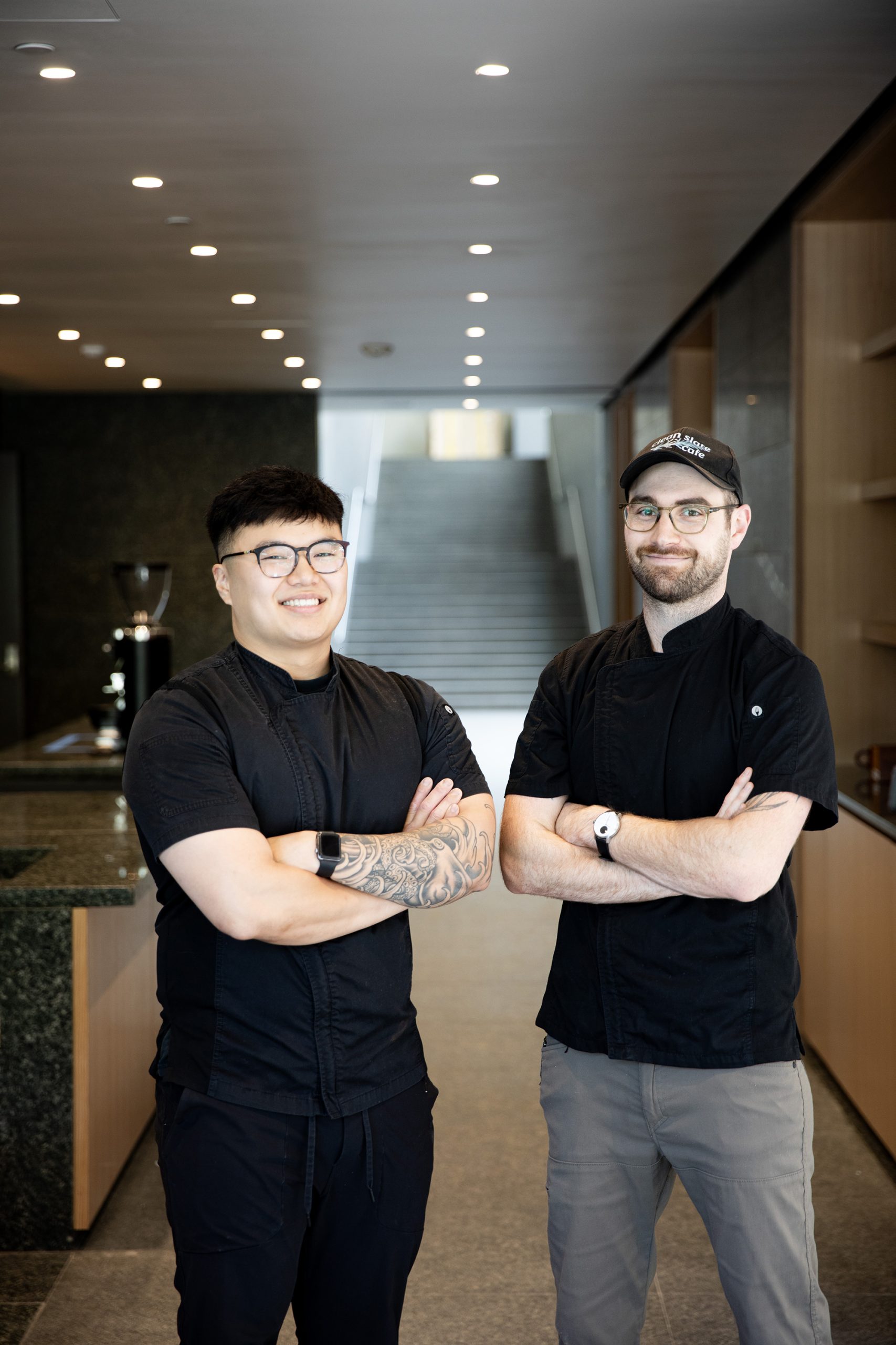 Two chefs stand side by side in a modern kitchen, smiling with arms crossed. The chef on the left has short black hair, glasses, tattoos on his arm, and wears a black outfit. The chef on the right wears a cap, glasses, and a black shirt with khaki pants.