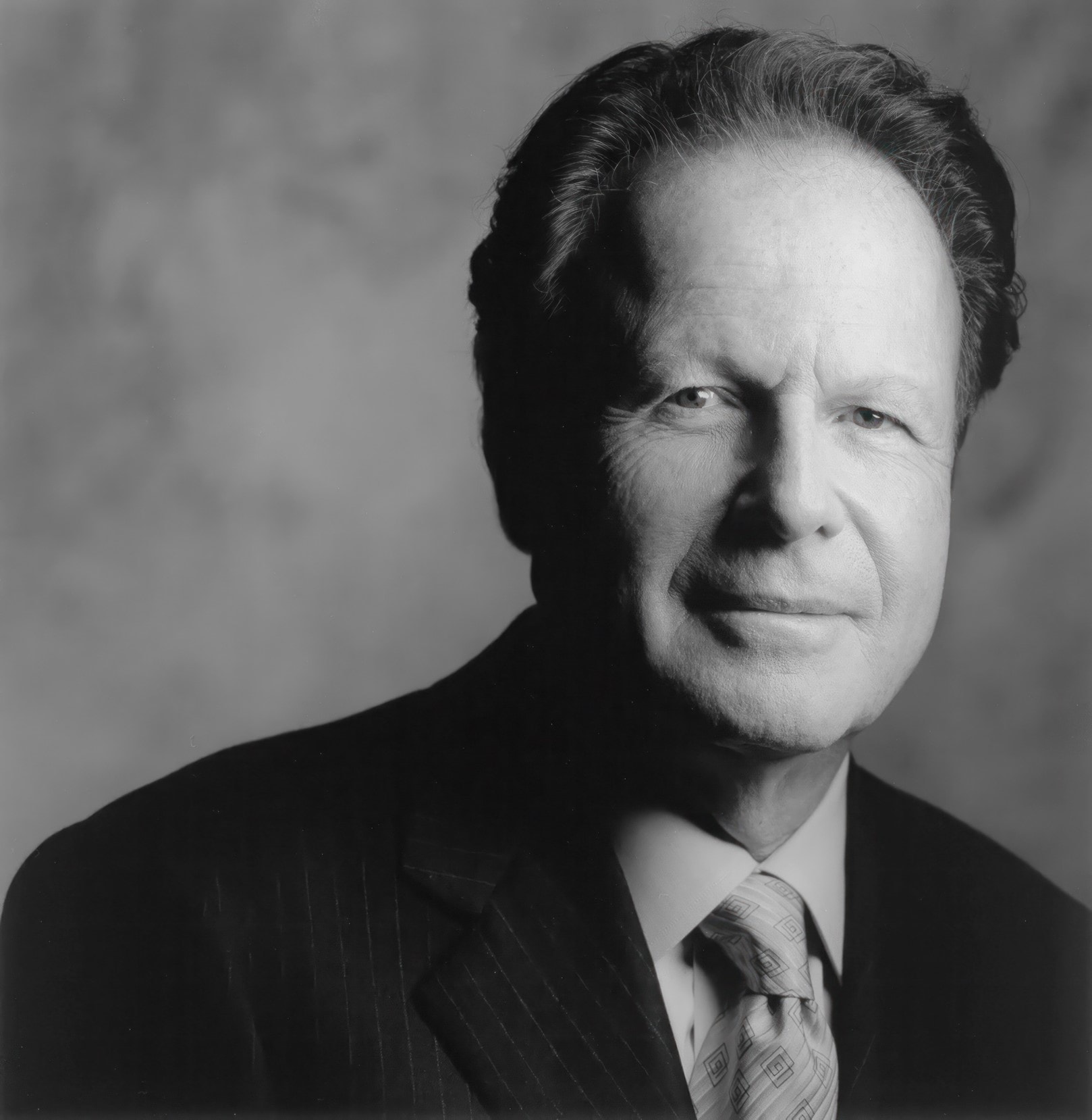 A black and white portrait of an older man with short, dark hair, dressed in a pinstripe suit and tie, looking directly at the camera with a neutral expression. The background is out-of-focus, creating a soft and professional ambiance.