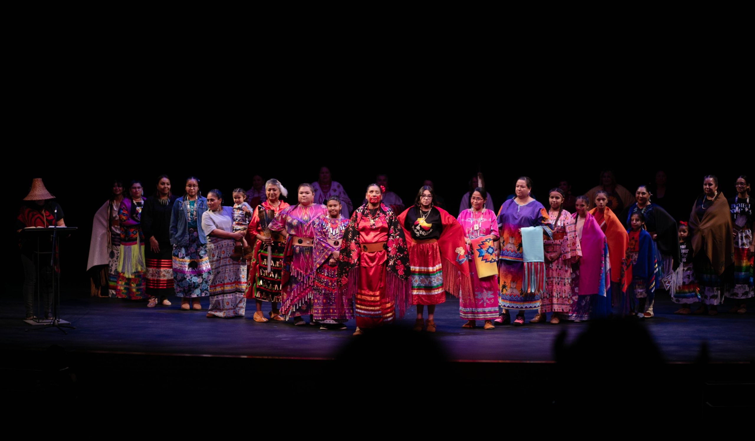 A diverse group of women dressed in vibrant, traditional attire stands in a line on a dimly lit stage, with each woman wearing a unique, colorful outfit. The backdrop is dark, highlighting the vivid colors of their clothing. A podium with a lamp is visible to the left.