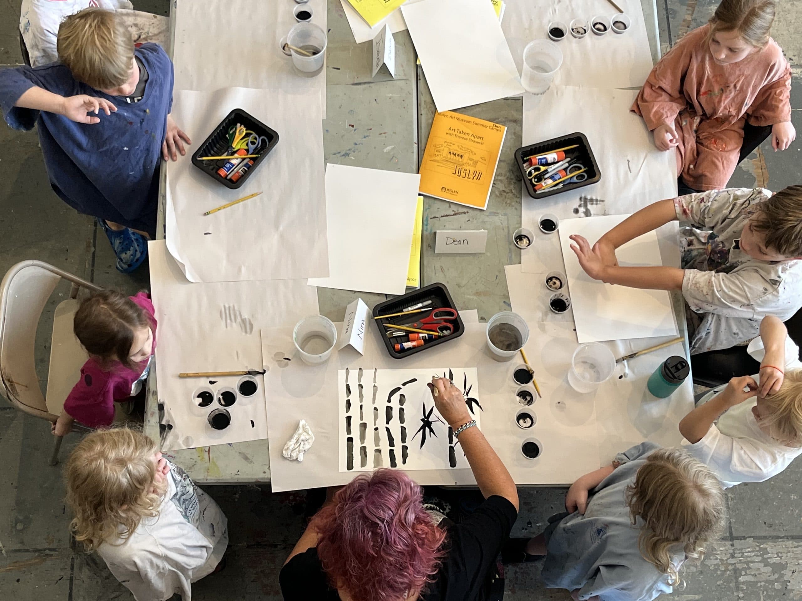 A group of children sit around a table as they are engaged in a painting activity. The table is covered with art supplies including brushes, containers of black paint, scissors, and papers with various designs. An adult supervises the activity.