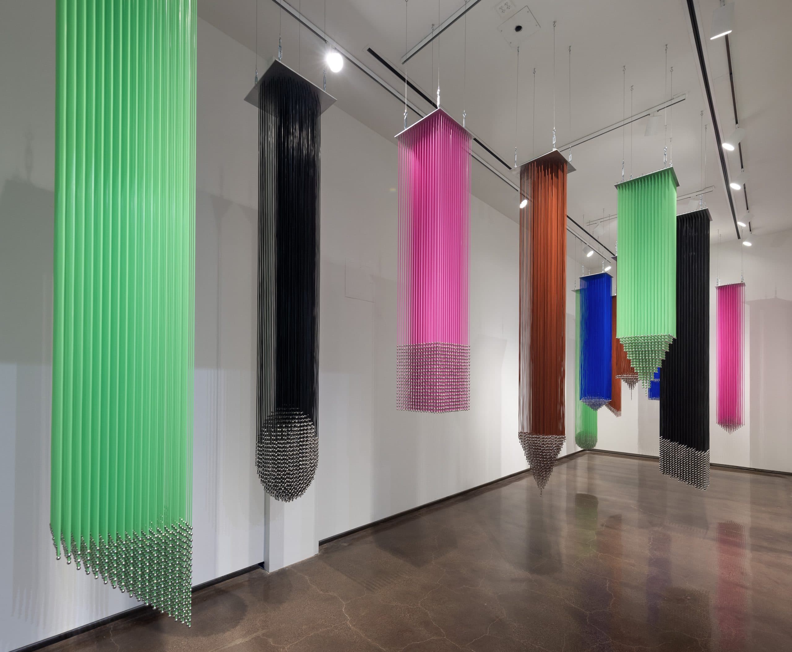 Hanging installations feature vertical tubes in various colors, including green, black, pink, brown, and blue, ending in pointed, bead-like tips. The artwork is displayed in a bright gallery with a polished floor.