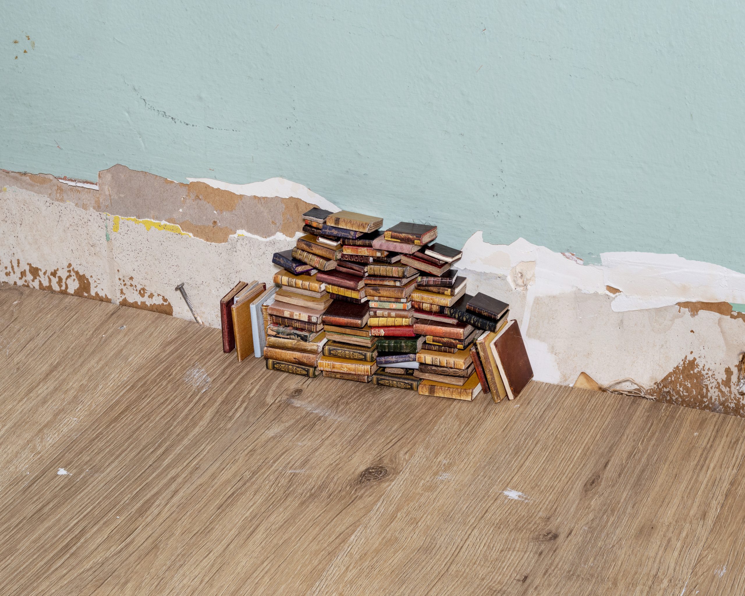 A small pyramid of miniature books stacked against a wall with peeling paint. The floor is light brown wood, contrasting with the wall's pale green color and the exposed layers underneath.