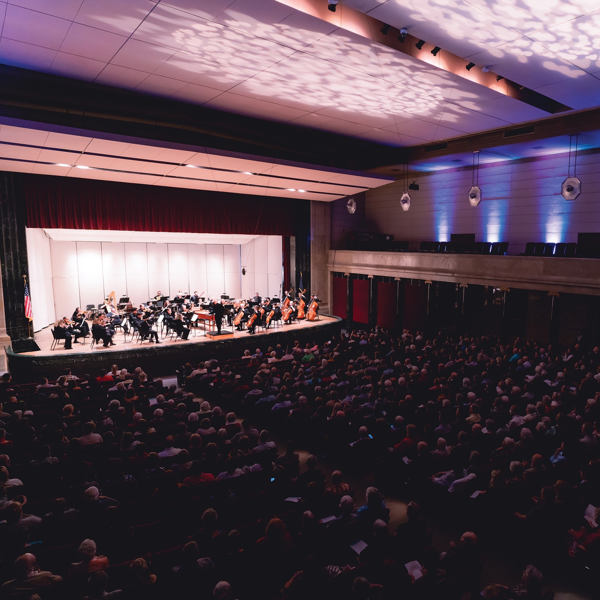 A symphony orchestra performs on a stage in a concert hall. The hall is filled with an audience seated in rows. The stage is illuminated with warm lights, and the ceiling features decorative lighting patterns.