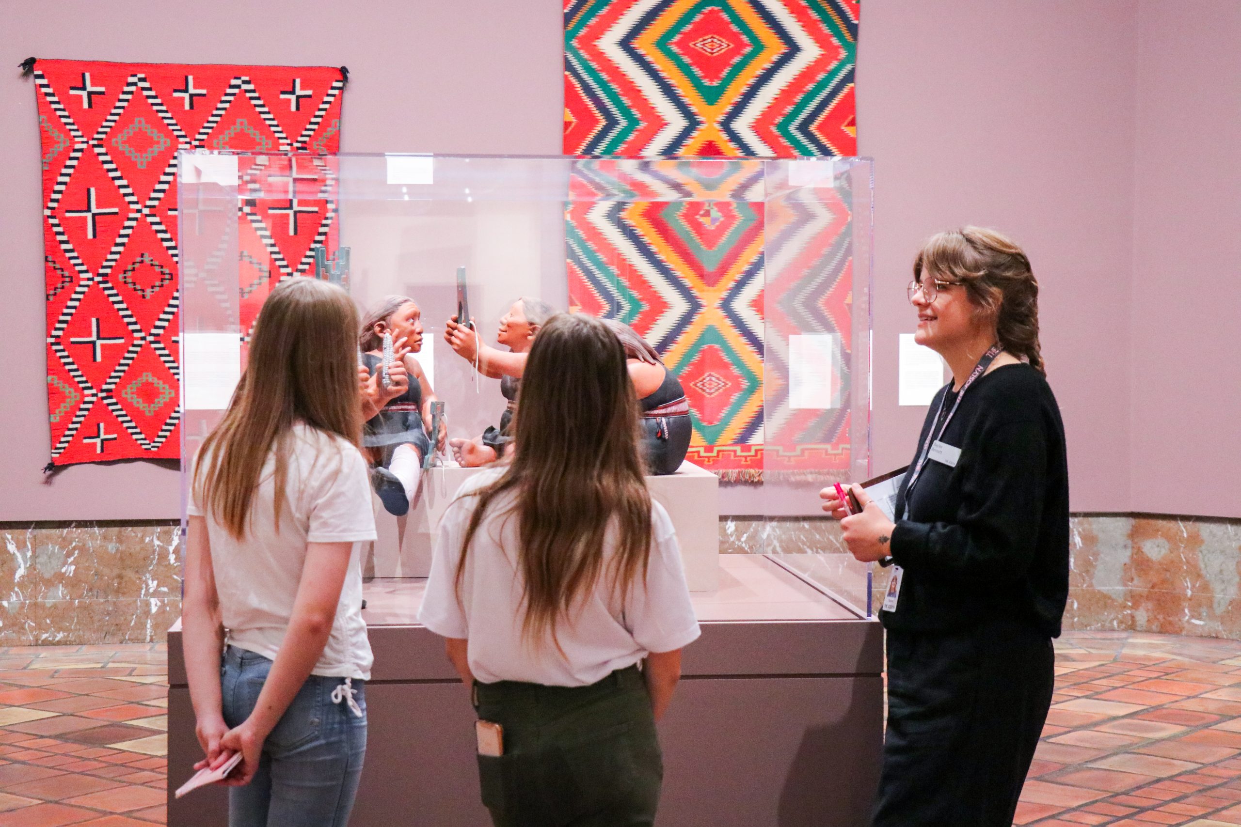 Three people view Native American pottery displayed in a museum case. Two have long hair and wear casual clothes. A woman in black attire explains the artwork. Colorful patterned tapestries are on the walls in the background.