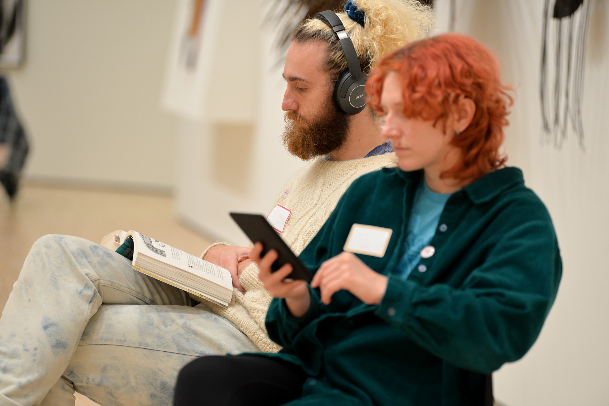 Two people sitting indoors. The person on the left is wearing headphones and reading a book, while the person on the right is using a smartphone. Both appear relaxed, possibly in a gallery or waiting area.